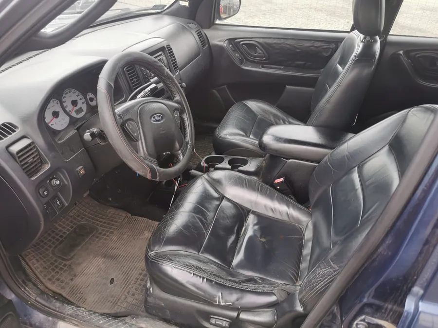 Wnętrze samochodu z widoczną czarną skórzaną tapicerką siedzeń w stanie zużytym z widocznymi pęknięciami. Kierownica z logo Ford, białe wskaźniki prędkościomierza z czerwonymi igłami, brudne dywaniki podłogowe oraz czarne paneli drzwiowe z teksturą. Widoczne są również uchwyty na napoje i fragment niebieskiego nadwozia po otwarciu drzwi.