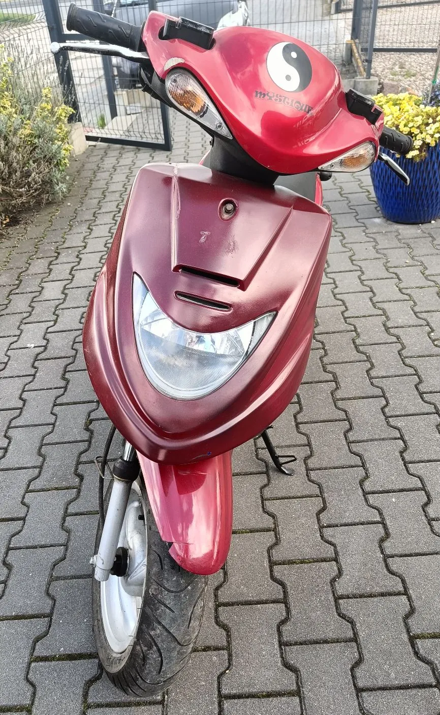 Czerwony skuter zaparkowany na szarej kostce brukowej. Na przedniej owiewce kierownicy znajduje się biało-czarny symbol yin-yang oraz napis 'MYSTIQUE'. Pojazd ma jeden duży przedni reflektor i dwa mniejsze kierunkowskazy po bokach kierownicy.