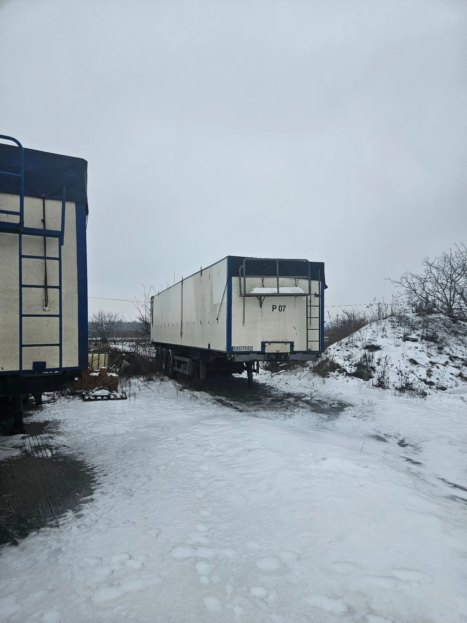 Zdjęcie przedstawia dwa białe pojazdy transportowe z niebieskimi obramowaniami, zaparkowane na zaśnieżonym terenie. Jeden z pojazdów znajduje się w centrum kadru, a drugi jest widoczny częściowo po lewej stronie. Na tylnej ścianie centralnego pojazdu widnieje napis 'P 07' oraz tablica rejestracyjna 'EKU FHF2'. W tle widać bezlistne krzewy i pagórek pokryty śniegiem pod zachmurzonym, szarym niebem.