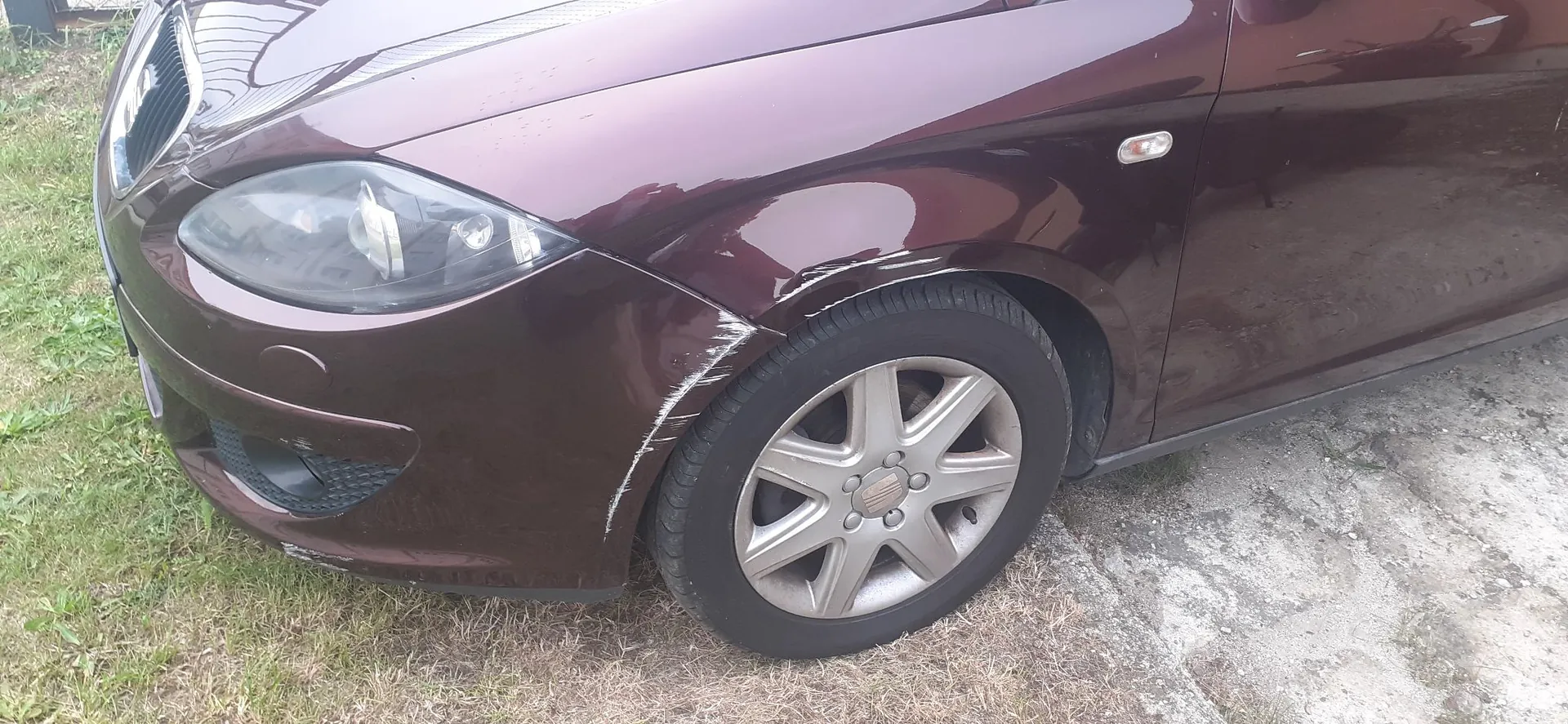 Zbliżenie na przedni prawy narożnik bordowego samochodu. Widoczne są uszkodzenia w postaci zarysowań i wgniecenia na zderzaku oraz błotniku nad przednim kołem z aluminianą felgą.