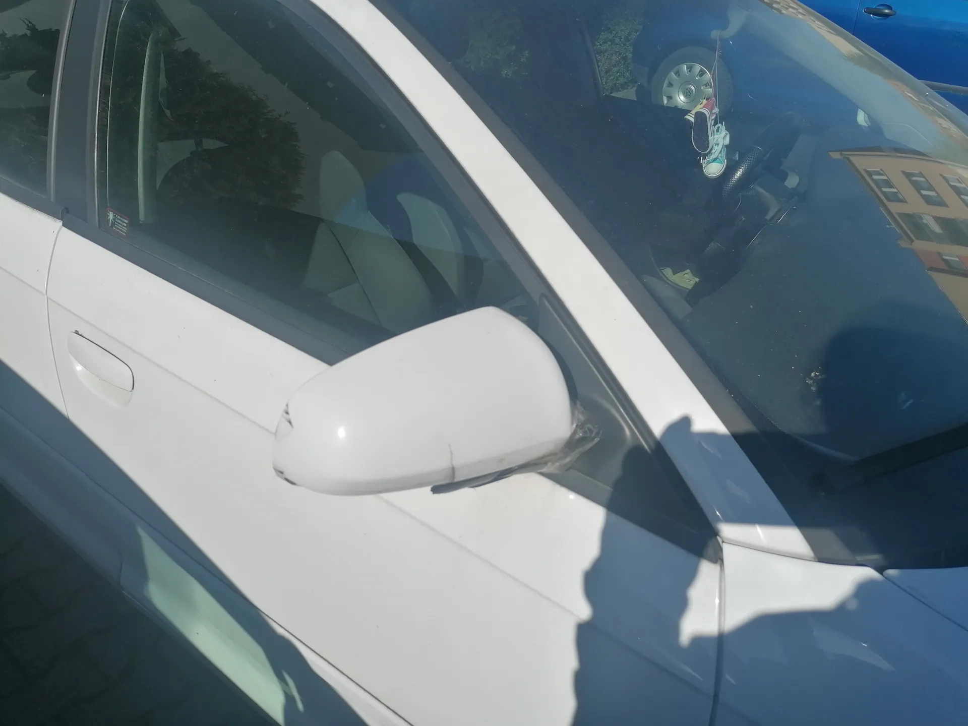Biały samochód widziany z boku, z lusterkiem bocznym owiniętym przezroczystą folią. W odbiciu na przedniej szybie widoczny niebieski samochód, budynek oraz cień osoby robiącej zdjęcie. Samochód zaparkowany na brukowanej powierzchni.