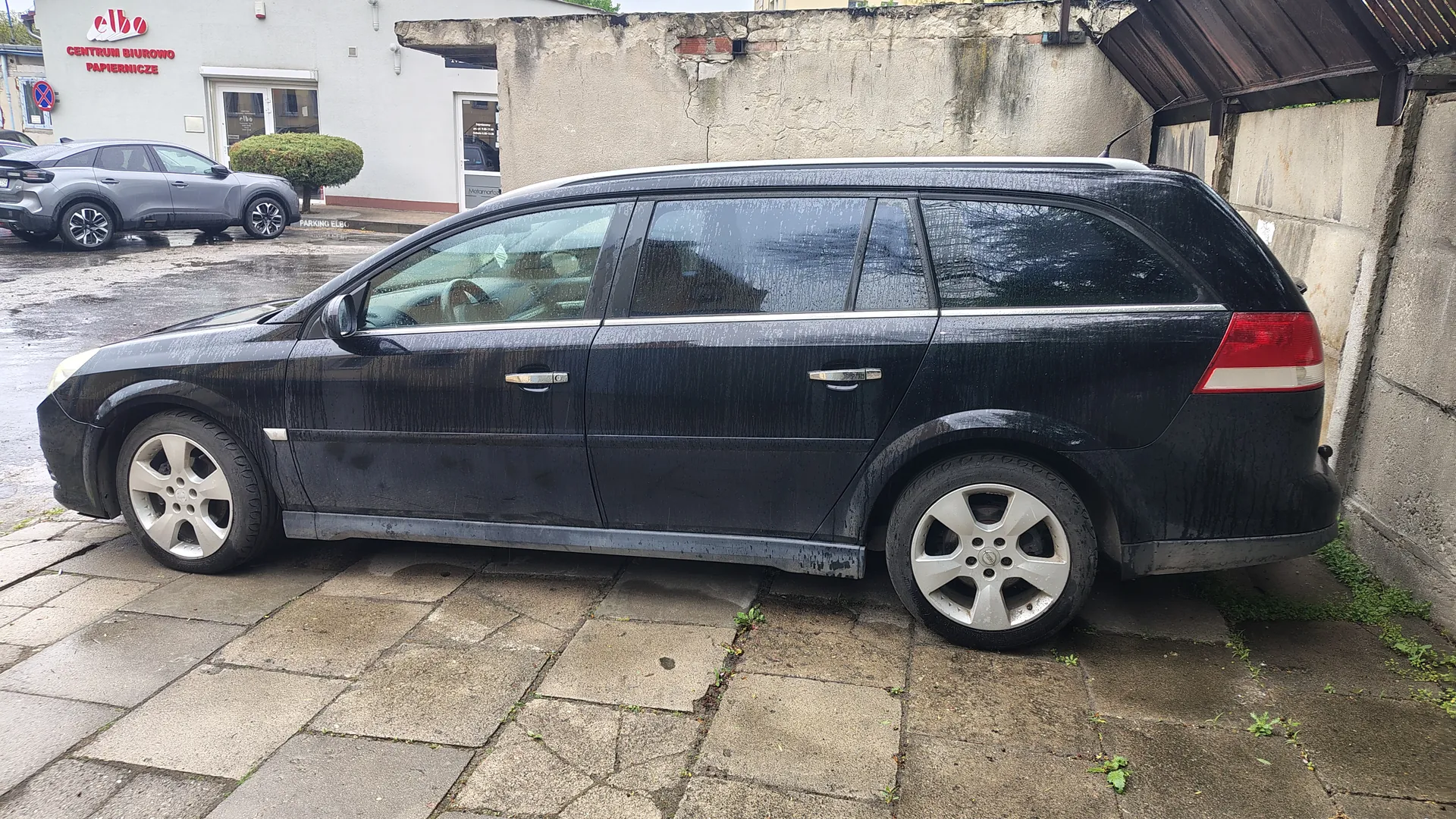 Czarny samochód kombi zaparkowany na brukowanej powierzchni, pokryty kroplami wody. W tle szary SUV, budynek z czerwonym napisem "elbo CENTRUM BIUROWO PAPIERNICZE" oraz tablicą "PARKING". Widoczna betonowa ściana, mokra nawierzchnia oraz zielone krzewy. Samochód ma srebrne felgi i ciemne szyby.
