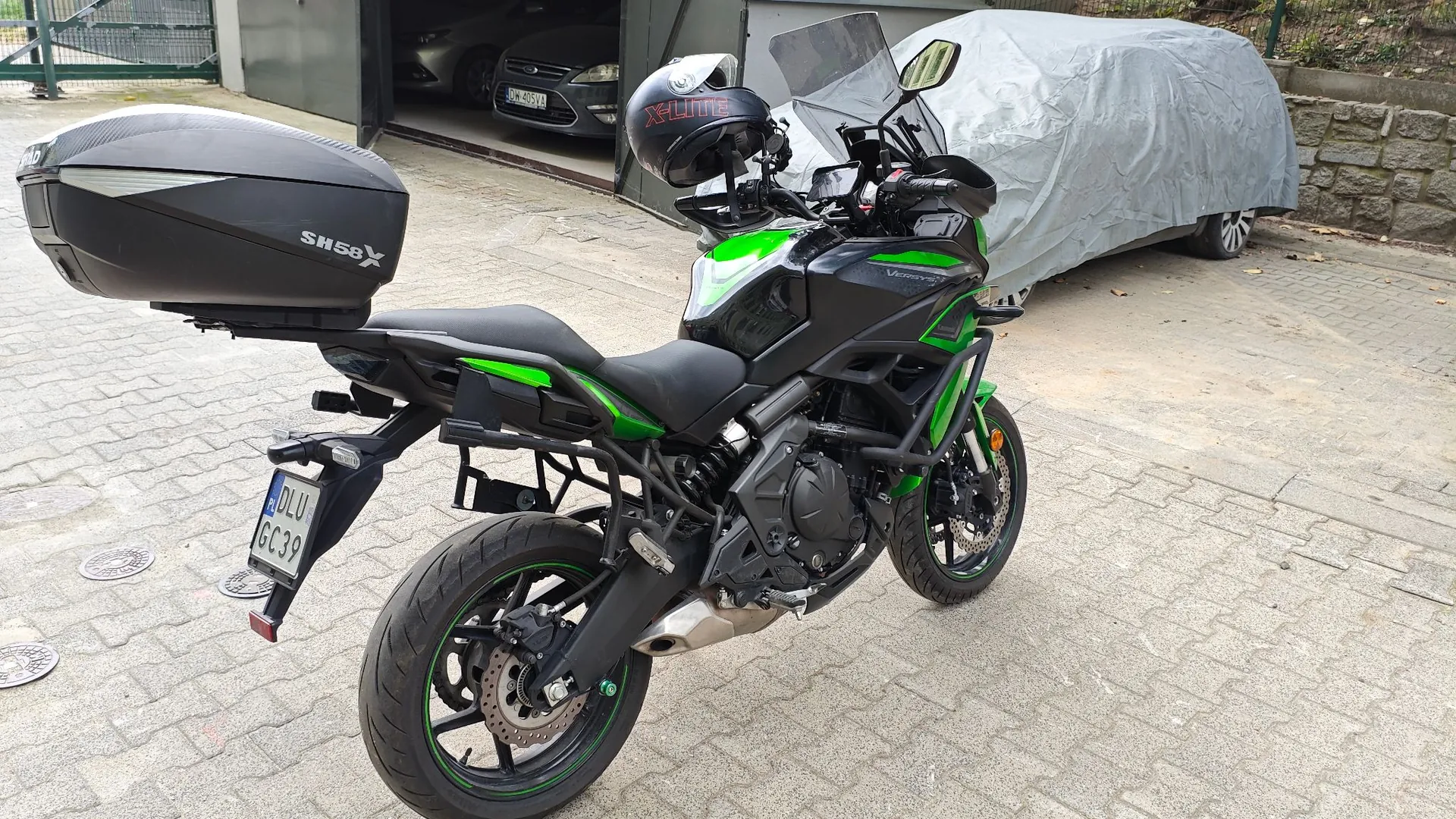 Czarny motocykl z zielonymi akcentami zaparkowany na kostce brukowej przed garażem. Motocykl posiada zamontowany czarny kufer tylny z napisem 'SH58X' oraz kask na lusterku. W tle widoczny jest otwarty garaż z samochodem oraz inny pojazd przykryty szarą plandeką. Tablica rejestracyjna motocykla to DLU GC39.