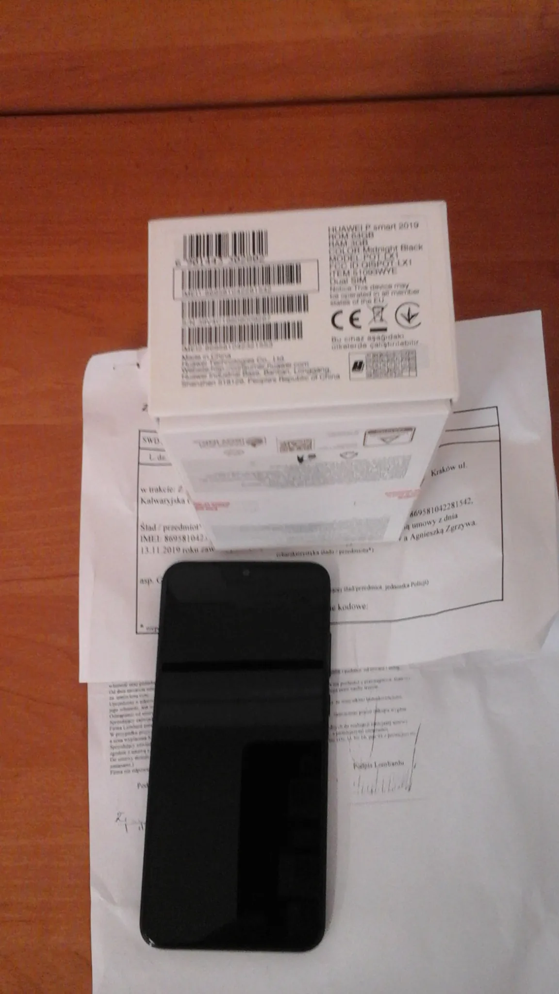 Czarny smartfon leżący na białych dokumentach z drukowanym tekstem, w tym widocznymi fragmentami 'Kodów ul.', '13.11.2019' oraz 'Podpis'. Obok znajduje się białe pudełko z napisem 'HUAWEI P smart 2019', kodami kreskowymi, znakiem CE i innymi oznaczeniami. Wszystko umieszczone na drewnianym blacie.