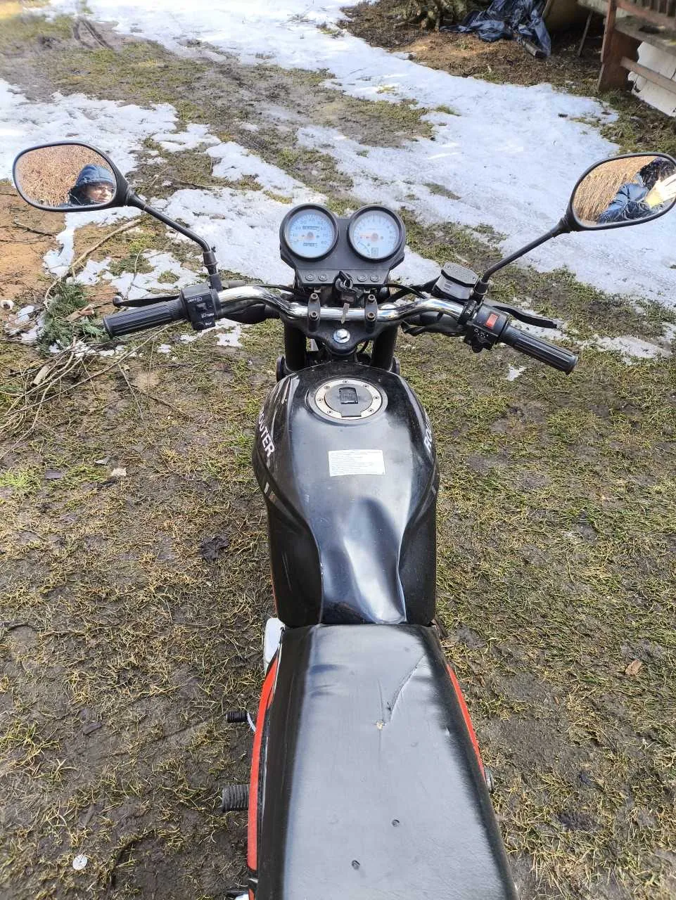 Czarny motocykl widziany z przodu, z widocznymi kierownicą, dwoma lusterkami bocznymi, dwoma wskaźnikami na desce rozdzielczej oraz bakiem paliwa z częściowo widocznym napisem HUNTER. W lewym lusterku odbicie osoby w niebieskiej kurtce z kapturem, w prawym lusterku odbicie słomy. Tło przedstawia ziemistą powierzchnię z resztkami śniegu i trawą, w oddali widoczne są czarne worki i fragment drewnianej konstrukcji.