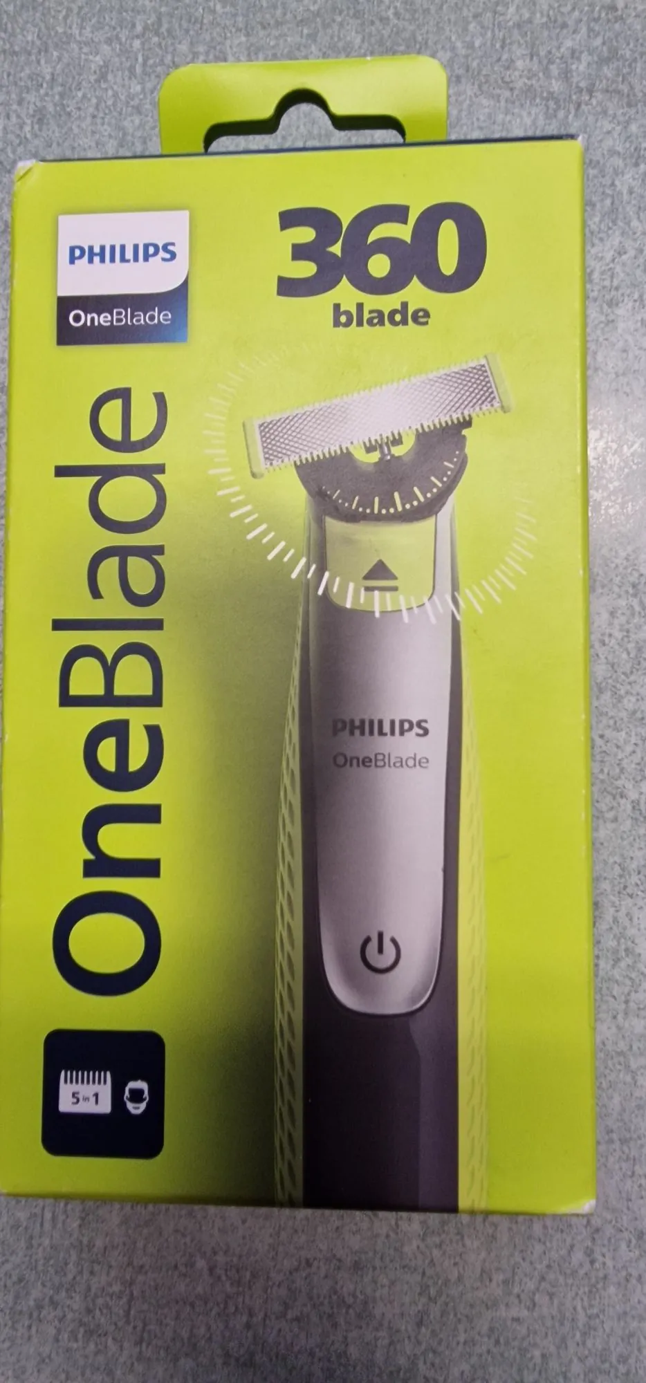 Opakowanie trymera Philips OneBlade. Pudełko jest w kolorze limonkowym z dużym napisem 'OneBlade' ułożonym pionowo po lewej stronie. W górnej części znajduje się napis '360 blade'. Na środku widnieje zdjęcie urządzenia z głowicą tnącą, na której naniesiono białą, przerywaną linię w kształcie półkola, sugerującą ruch obrotowy. Na obudowie urządzenia widnieje logo 'PHILIPS OneBlade' oraz przycisk zasilania. W lewym dolnym rogu znajduje się czarna ikona z napisem '5 in 1'.