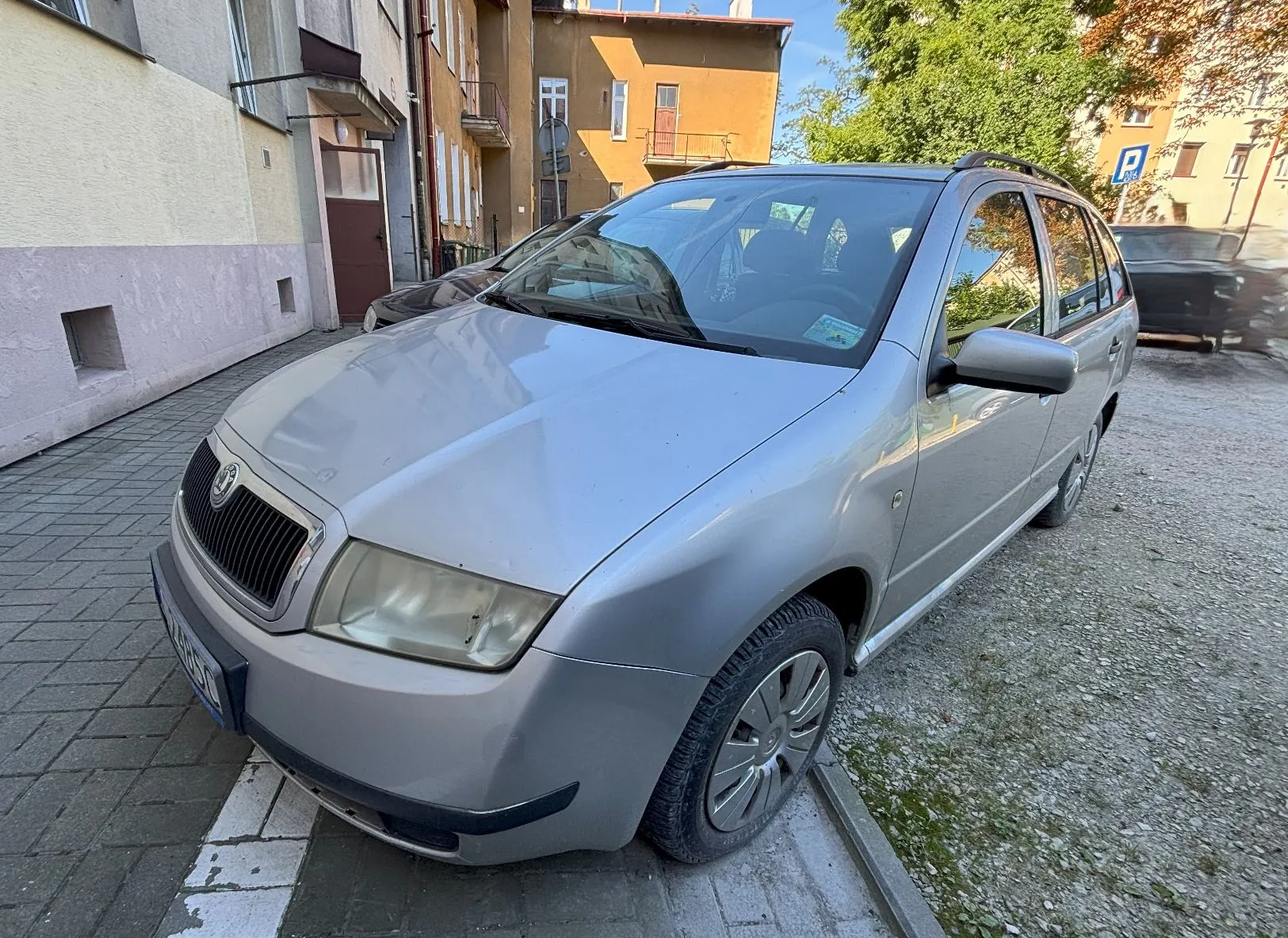 Srebrny samochód marki Skoda w nadwoziu kombi, zaparkowany na kostce brukowej przed budynkami mieszkalnymi. Pojazd jest widoczny z przodu i z boku, z lekkim kątem. W tle widać jasne elewacje budynków oraz zielone drzewa.