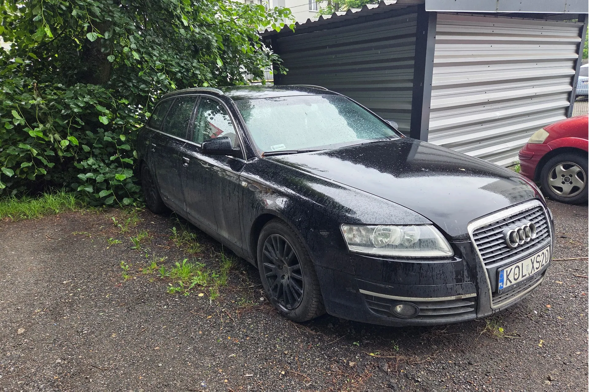 Czarny samochód marki Audi w nadwoziu kombi, zaparkowany na żwirowym podjeździe obok metalowego garażu i zielonych krzewów. Samochód jest brudny i mokry od deszczu. Widoczna tablica rejestracyjna o treści KOL XS20. W tle po prawej stronie widoczny jest fragment czerwonego samochodu.