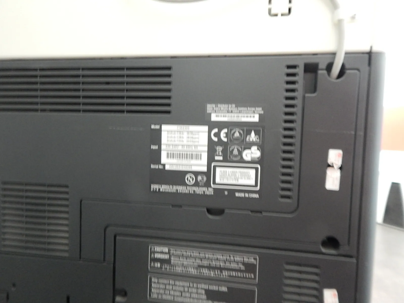 Zdjęcie tylnej obudowy czarnego urządzenia elektronicznego, prawdopodobnie monitora lub telewizora. Widoczne są otwory wentylacyjne w formie poziomych i pionowych żeberek oraz srebrna naklejka z danymi technicznymi, certyfikatami (m.in. CE) i kodem kreskowym. Na dole znajduje się kolejna naklejka z ostrzeżeniami w języku angielskim i niemieckim. W prawym górnym rogu widoczny jest biały kabel zasilający.