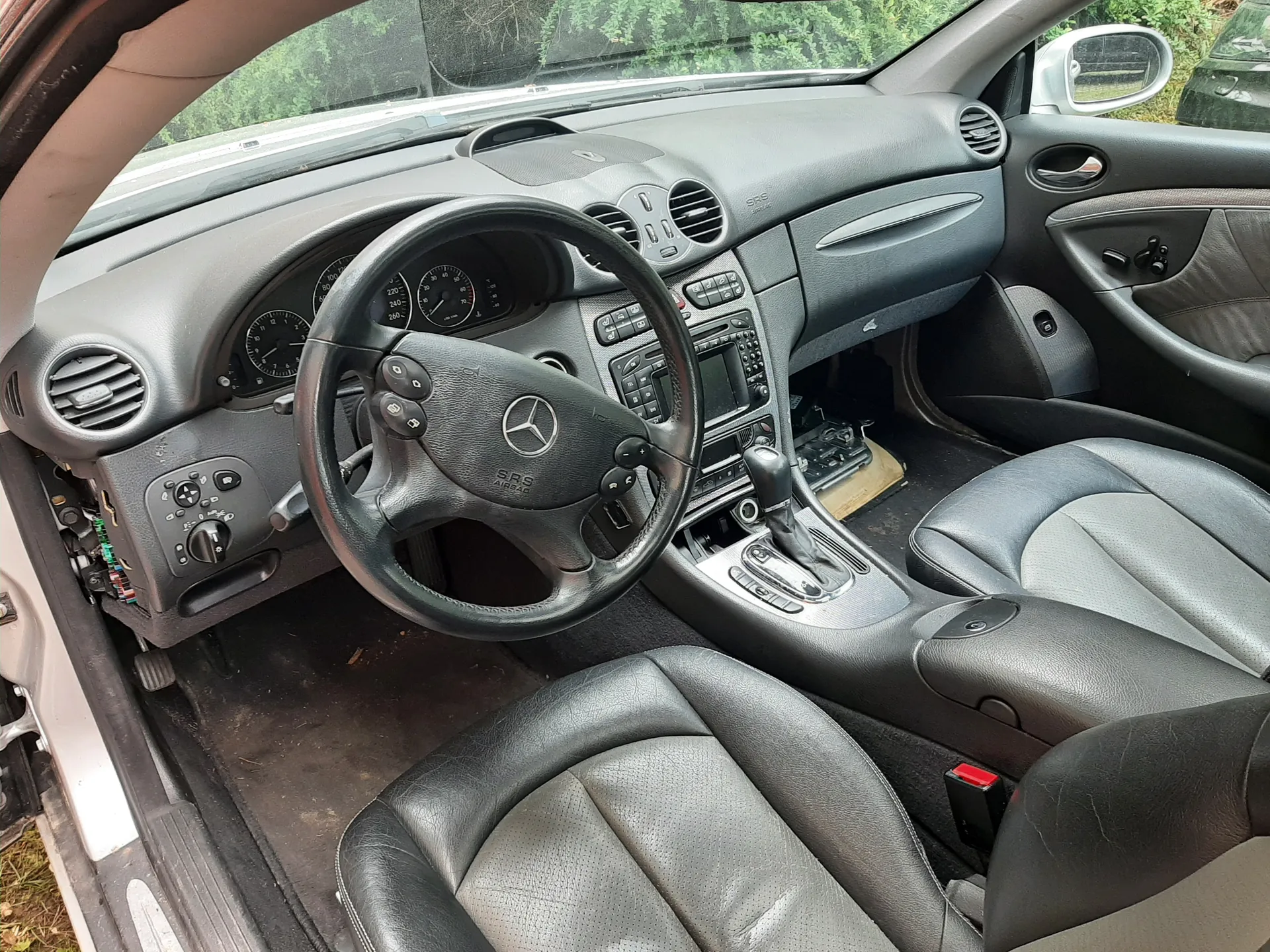 Wnętrze samochodu Mercedes-Benz z kierownicą z logo i napisem SRS AIRBAG, szarą deską rozdzielczą z licznikami, przyciskami i centralnym panelem sterowania, czarnymi skórzonymi fotelami, otwartymi drzwiami kierowcy oraz widokiem na zieloną roślinność przez przednią szybę. Po lewej stronie deski rozdzielczej widoczne są odsłonięte przewody elektryczne.