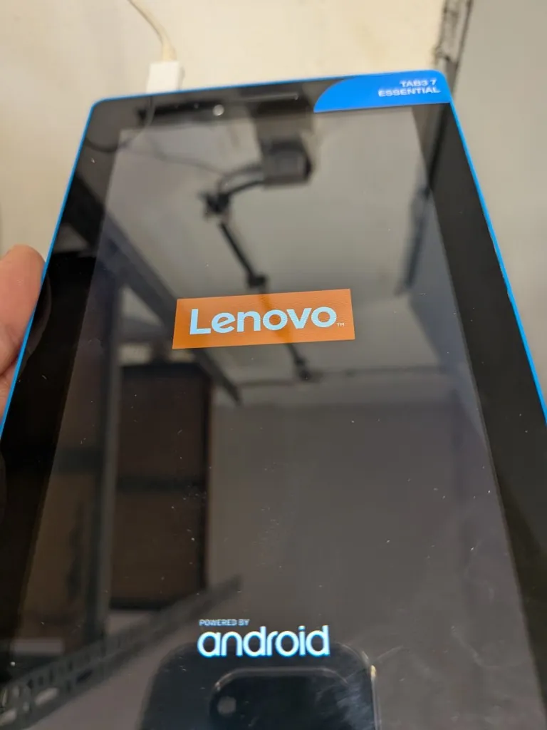 Zdjęcie tabletu Lenovo Tab3 7 Essential z przodu. Urządzenie ma czarny ekran z niebieskimi ramkami po bokach. Na środku ekranu znajduje się pomarańczowy prostokąt z białym napisem „Lenovo”, a na górnej krawędzi ramki widnieje napis „TAB3 7 ESSENTIAL”. Na dole ekranu znajduje się biały napis „powered by android”. W lewym górnym rogu widoczny jest fragment palca osoby trzymającej urządzenie.