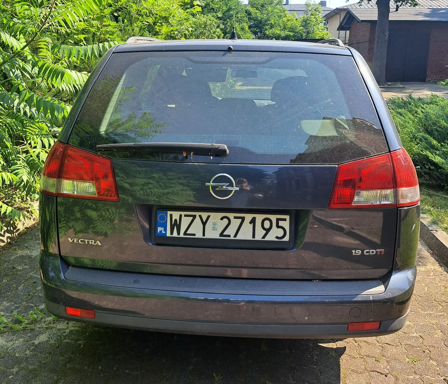 Tylny widok ciemnoniebieskiego samochodu marki Opel Vectra 1.9 CDTI. Pojazd posiada polską tablicę rejestracyjną o numerze WZY 27195. Samochód zaparkowany jest na podjeździe w otoczeniu zielonych drzew i krzewów.