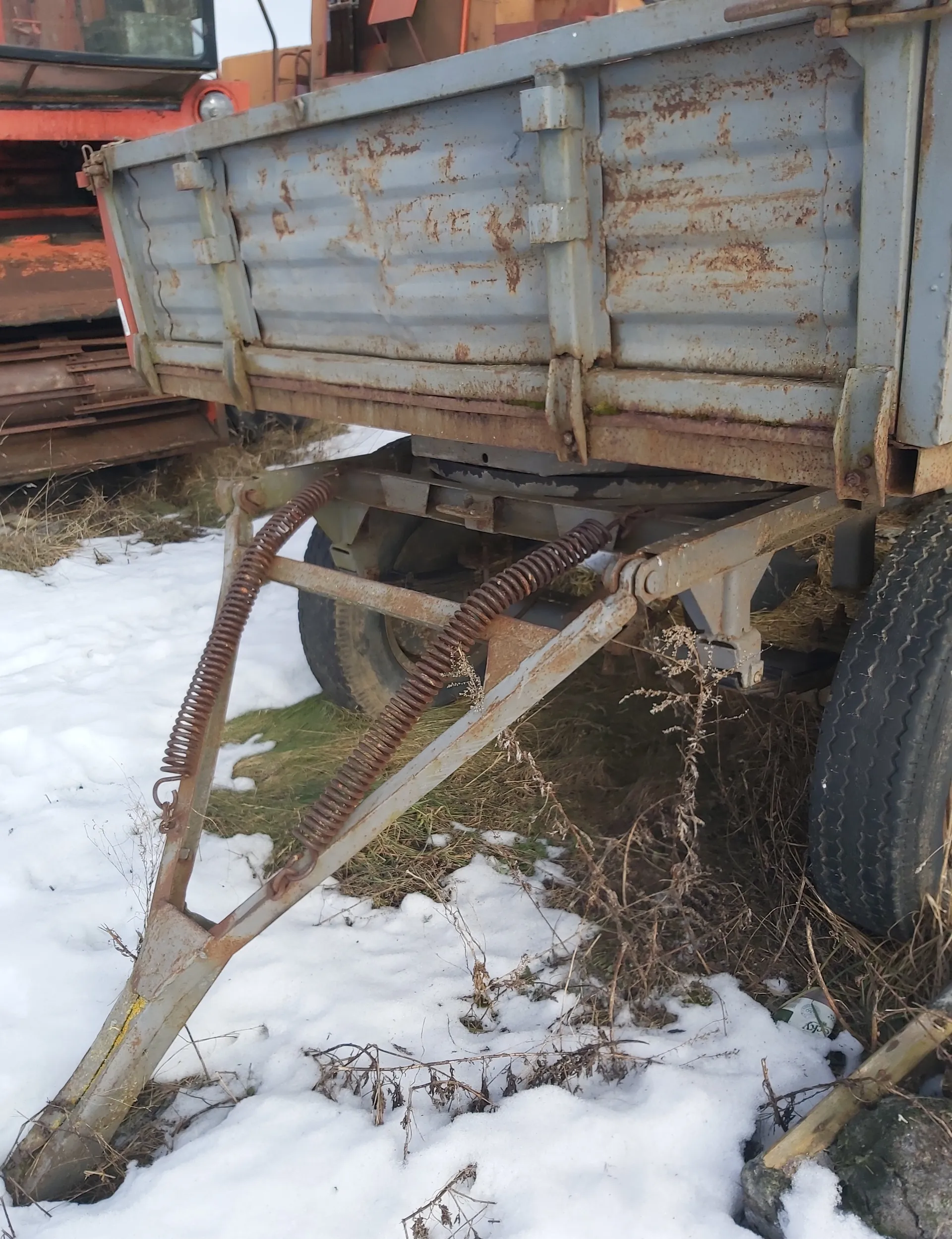 Zardzewiały metalowy przyczepa lub tylna część pojazdu z widocznymi sprężynami i metalowymi wspornikami. Na tle pomarańczowy pojazd rolniczy lub budowlany. Podłoże pokryte śniegiem i suchą trawą. Widoczne opony oraz zardzewiałe elementy konstrukcyjne.