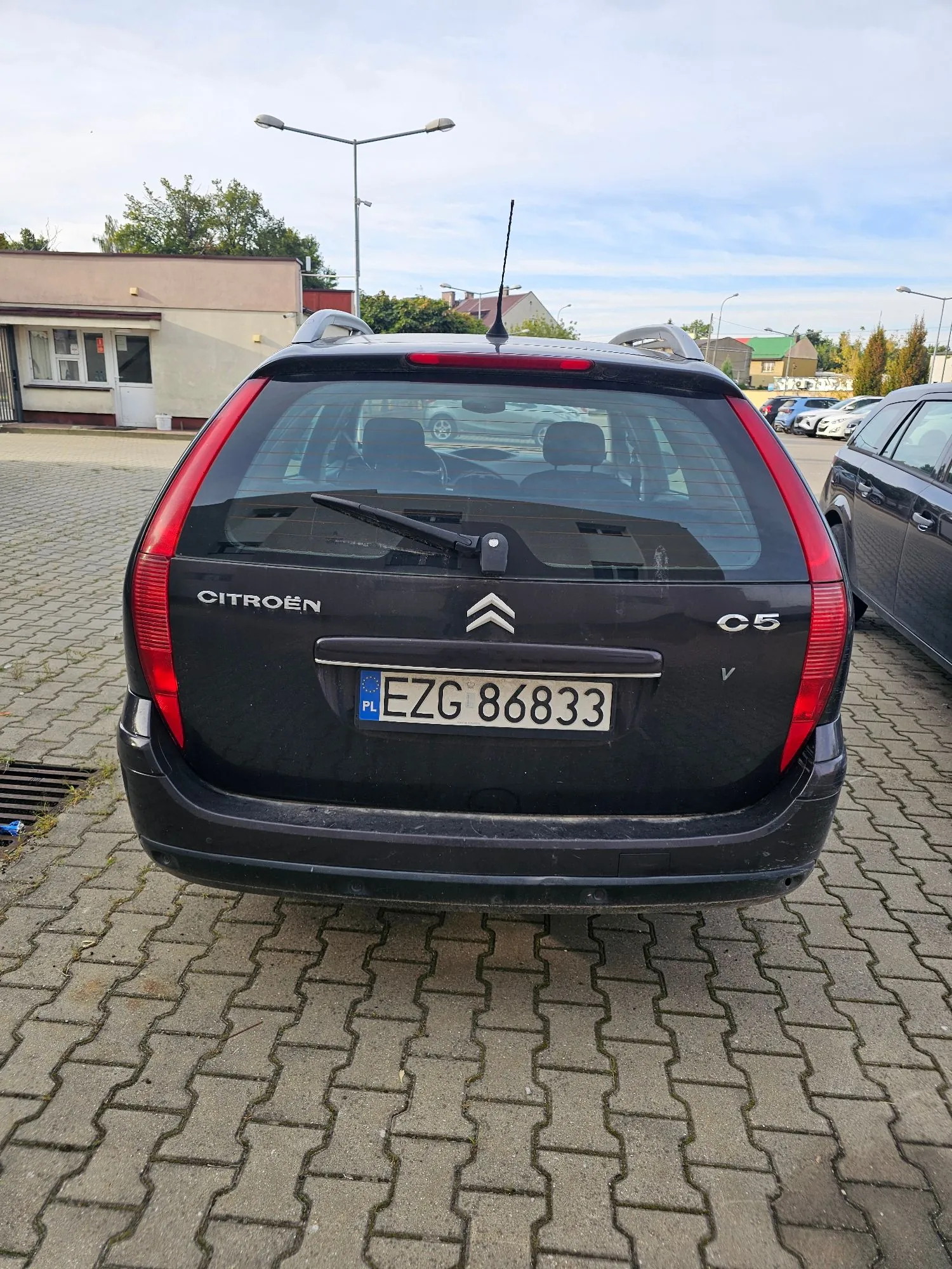 Tylny widok czarnego samochodu marki Citroën model C5 zaparkowanego na kostce brukowej. Na klapie bagażnika widoczne jest logo marki, napis CITROËN, oznaczenie modelu C5 oraz polska tablica rejestracyjna o numerze EZG 86833. W tle widać niskie budynki, latarnie uliczne i inne zaparkowane samochody pod jasnym, lekko zachmurzonym niebem.