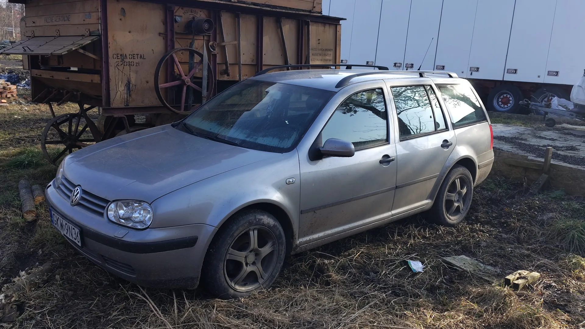 Srebrny samochód osobowy typu kombi marki Volkswagen z tablicą rejestracyjną GKW 9N36, zaparkowany na trawiastym terenie. W tle drewniany przyczepa z napisami CENA DETAL 27000 zl i PALENE WZOROWE oraz biały naczepa ciężarowa. Na ziemi widoczne są kawałki drewna i śmieci.