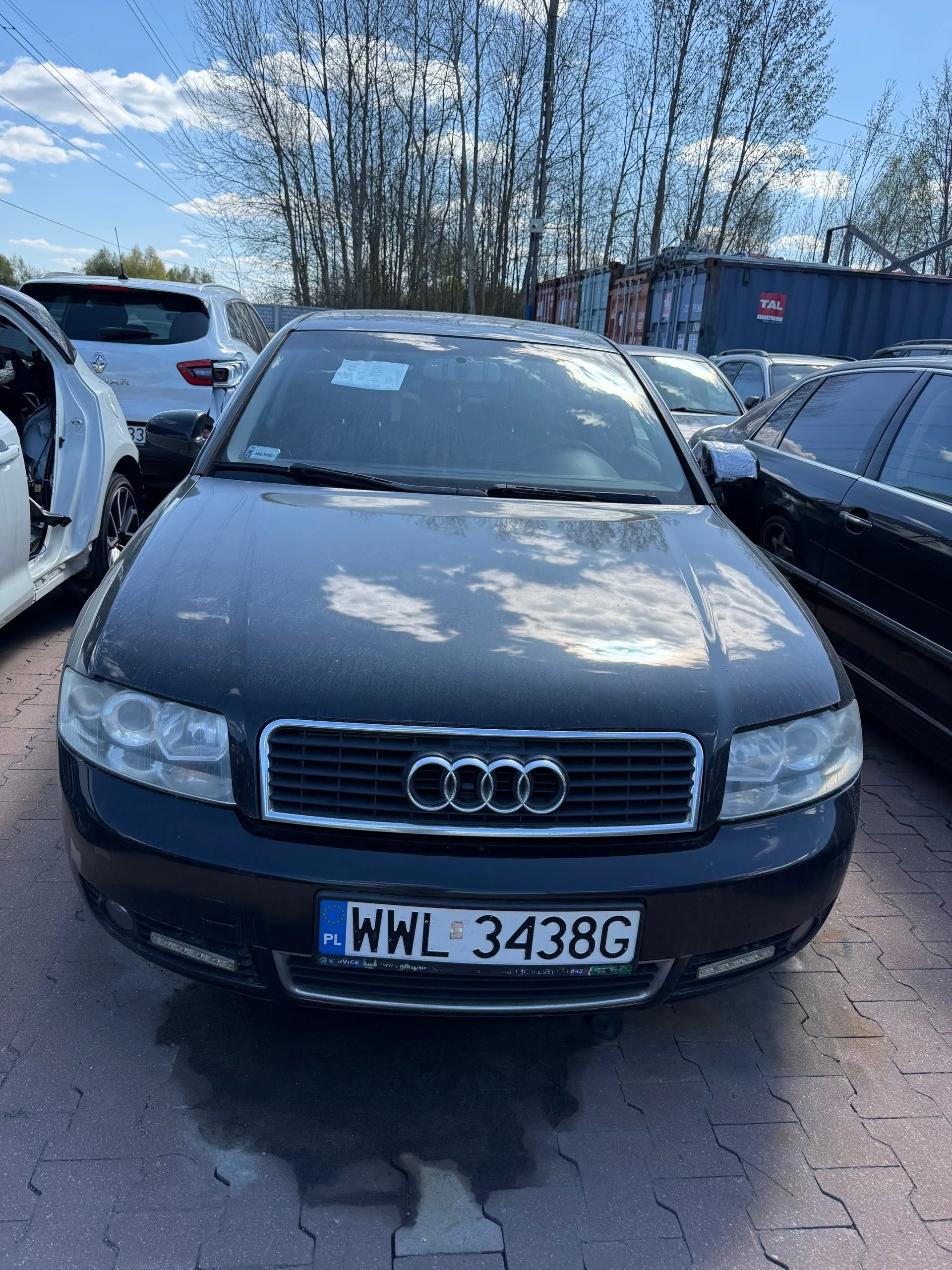 Czarny samochód marki Audi z przodu, z widoczną tablicą rejestracyjną PL WWL 3438G. Po lewej stronie biały samochód z otwartą drzwią, po prawej czarny samochód. W tle drzewa bez liści, kontenery (w tym z napisem TAL), niebo z chmurami. Samochody stoją na brukowanej powierzchni.