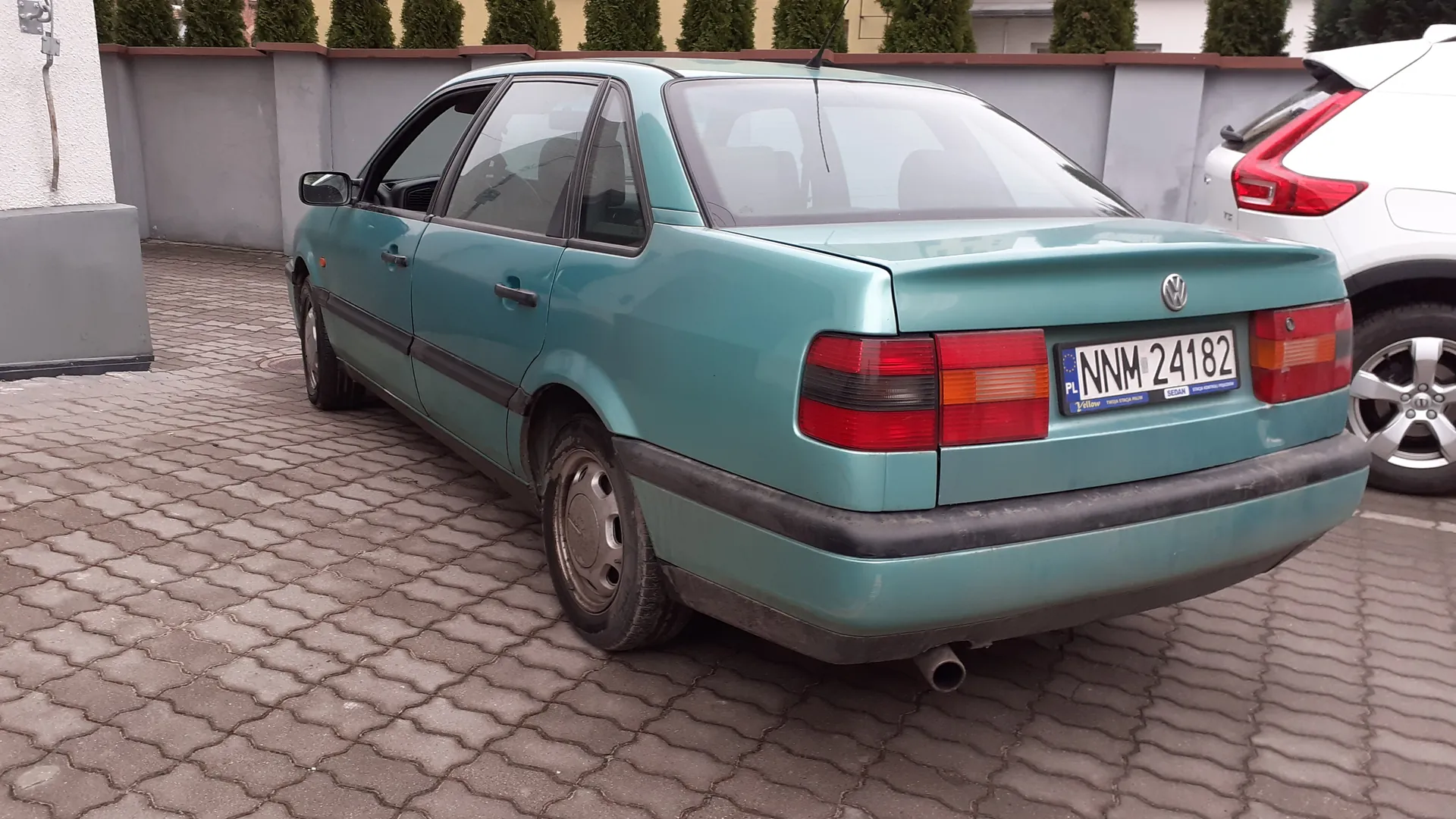 Zdjęcie przedstawia tył turkusowego samochodu osobowego marki Volkswagen, zaparkowanego na brukowanej powierzchni. Widoczna jest tablica rejestracyjna z polskim kodem PL i numerem NNM 24182, a także fragment białego samochodu po prawej stronie. W tle znajduje się szara ściana z brązowym wykończeniem oraz zielone krzewy. Samochód ma czarne zderzaki i czerwone światła tylnego. Tekst na tablicy rejestracyjnej jest czytelny i poziomo ustawiony.