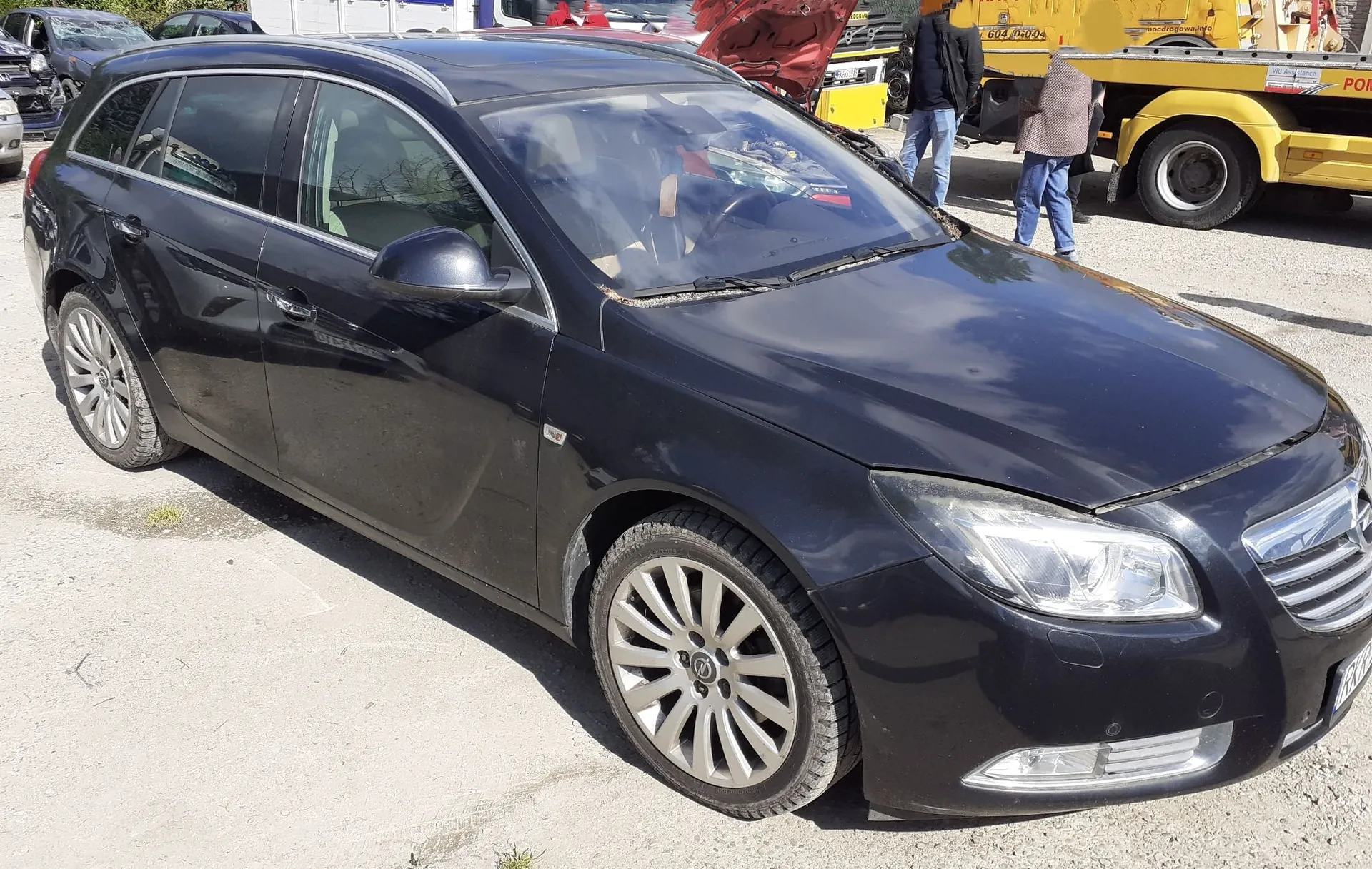 Czarny samochód typu kombi zaparkowany na betonowym placu. Pojazd jest zakurzony, a na dachu widoczne są uszkodzenia w postaci wgnieceń i odkształceń blachy. W tle widoczny jest żółty samochód laweta oraz dwie osoby stojące obok niej.