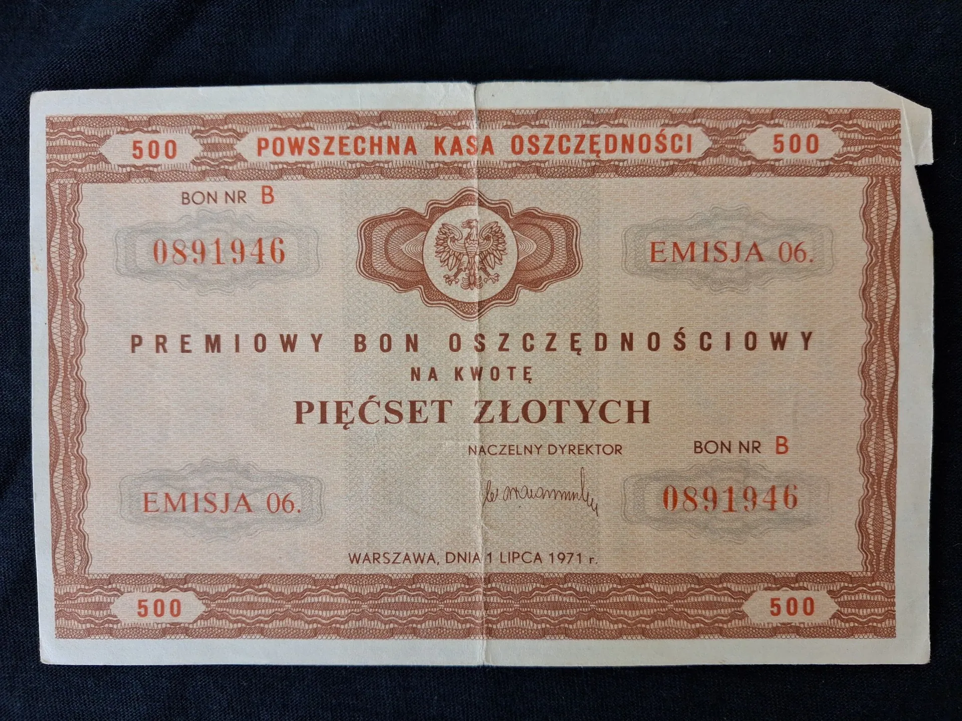Zdjęcie starego, papierowego dokumentu o nazwie 'PREMIOWY BON OSZCZĘDNOŚCIOWY' wydanego przez Powszechną Kasę Oszczędności. Dokument ma brązowe zdobienia w formie giloszy na krawędziach i w centrum znajduje się godło Polski. Tekst na dokumencie zawiera: 'POWSZECHNA KASA OSZCZĘDNOŚCI', nominał '500' w czterech rogach, numer bonu 'B 0891946' powtórzony dwukrotnie, napis 'EMISJA 06.' powtórzony dwukrotnie oraz kwotę 'PIĘĆSET ZŁOTYCH'. Na dole widnieje napis 'WARSZAWA, DNIA 1 LIPCA 1971 r.' oraz podpis Naczelnego Dyrektora.