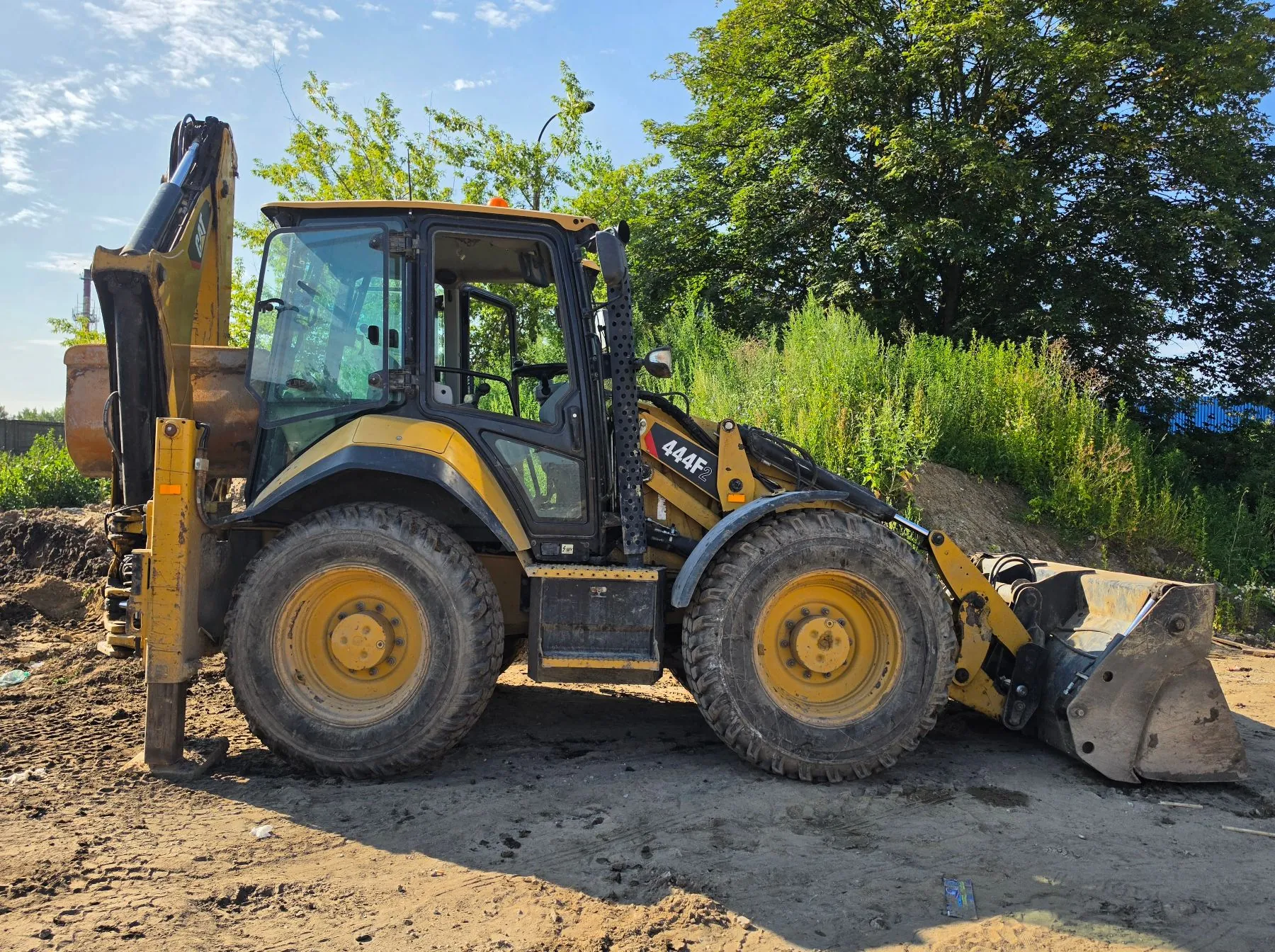 Żółta koparko-ładowarka marki JCB, model 444F, zaparkowana na piaszczystym placu budowy. Maszyna jest widoczna z profilu, z dużym ładowaczem z przodu i ramieniem koparki z tyłu. W tle znajdują się zielone drzewa i krzewy pod jasnoniebieskim niebem z lekkimi chmurami.