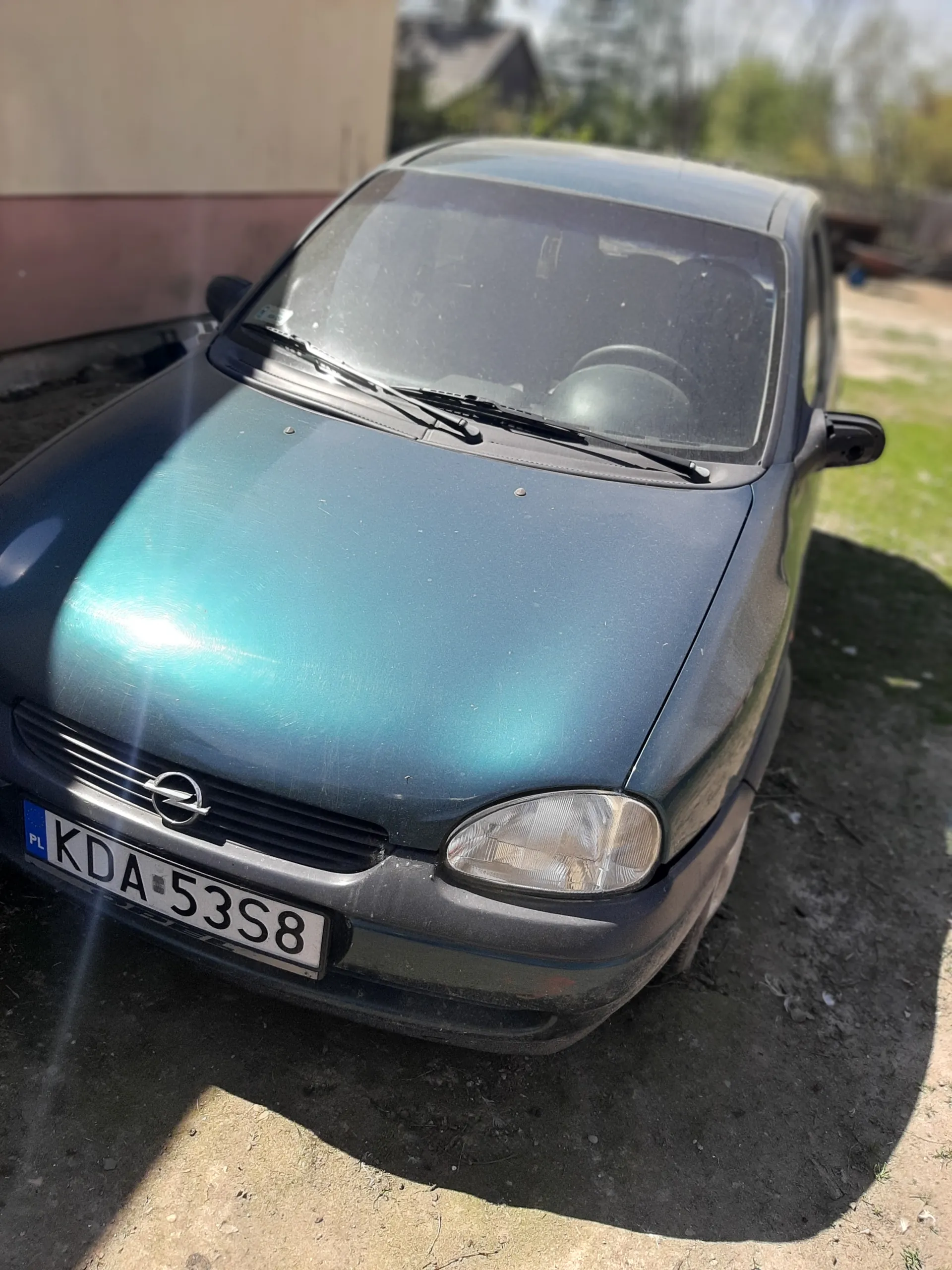 Niebieski samochód marki Opel, widok z przodu, tablica rejestracyjna KDA 5338 z niebieskim pasem PL po lewej stronie. Samochód zaparkowany na nawierzchni asfaltowej, w tle trawa i fragment budynku. Widoczne przednie światło oraz wycieraczki na przedniej szybie.