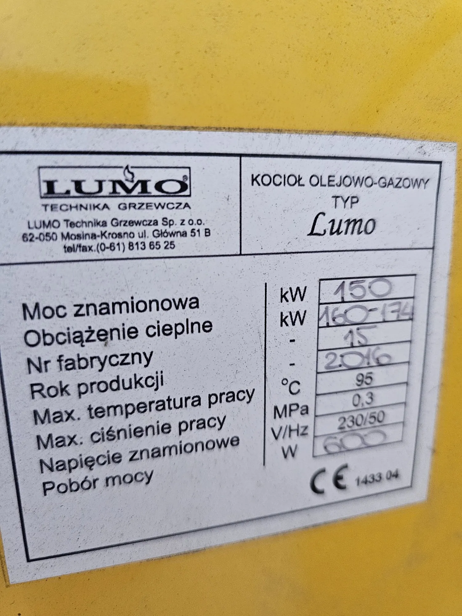 Zdjęcie tabliczki znamionowej urządzenia. W górnej części znajduje się logo 'LUMO TECHNIKA GRZEWCZA' oraz dane firmy: 'LUMO Technika Grzewcza Sp. z o.o. 62-050 Mosina-Krosno ul. Główna 51 B tel/fax.(0-61) 813 65 25'. Obok widnieje napis 'KOCIOŁ OLEJOWO-GAZOWY TYP Lumo'. Poniżej znajduje się tabela z parametrami technicznymi: Moc znamionowa: 150 kW, Obciążenie cieplne: 160-174 kW, Nr fabryczny: 15, Rok produkcji: 2016, Max. temperatura pracy: 95 °C, Max. ciśnienie pracy: 0,3 MPa, Napięcie znamionowe: 230/50 V/Hz, Pobór mocy: 600 W. W prawym dolnym rogu widnieje znak CE oraz numer 1433 04.