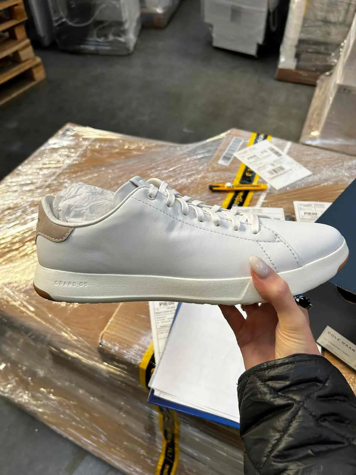 Biała trampka z brązowym akcentem na pięcie, trzymana w dłoni z jasnym manicurem. Na podeszwie widoczny napis 'GRAND.OS'. W tle: opakowania owinięte folią, żółty nożyk, dokumenty z częściowo czytelnym tekstem 'PIE' oraz palety w magazynie. Osoba trzymająca but ubrana jest w czarną pikowaną kurtkę.