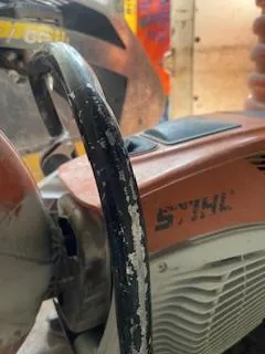 Zbliżenie na pomarańczowe urządzenie marki STIHL z czarnym uchwytem pokrytym odłupaną białą farbą. Widoczny czarny napis STIHL na boku urządzenia. W tle rozmyte elementy warsztatowe, w tym drewniane deski i czerwony przedmiot.