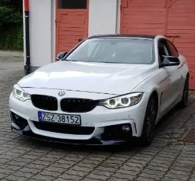 Biały samochód marki BMW zaparkowany na kostce brukowej przed budynkiem z czerwonymi drzwiami. Pojazd posiada czarne detale, w tym felgi i przedni spojler. Tablica rejestracyjna samochodu to ZSZ 38152.