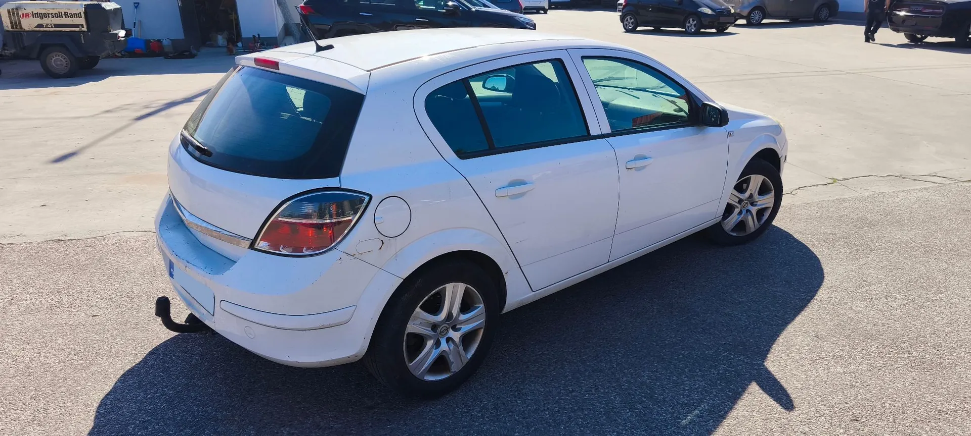 Biały samochód osobowy marki Opel Astra w nadwoziu hatchback, zaparkowany na betonowym placu. Pojazd jest pokazany z perspektywy tylno-bocznej od strony pasażera. W tle widoczne są inne zaparkowane samochody oraz fragmenty budynków przemysłowych, w tym maszyna z napisem 'Ingersoll Rand'.