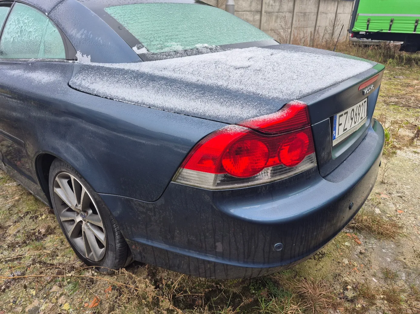 Tylne widok ciemnoniebieskiego samochodu z lekkim pokryciem śniegu na bagażniku. Widoczna tablica rejestracyjna PL FZ 9692T. Czerwone światła tylnego oświetlenia, błotne plamy na karoserii. W tle zielona naczepa ciężarówki i betonowa ściana. Podłoga z błotem i suchą trawą.