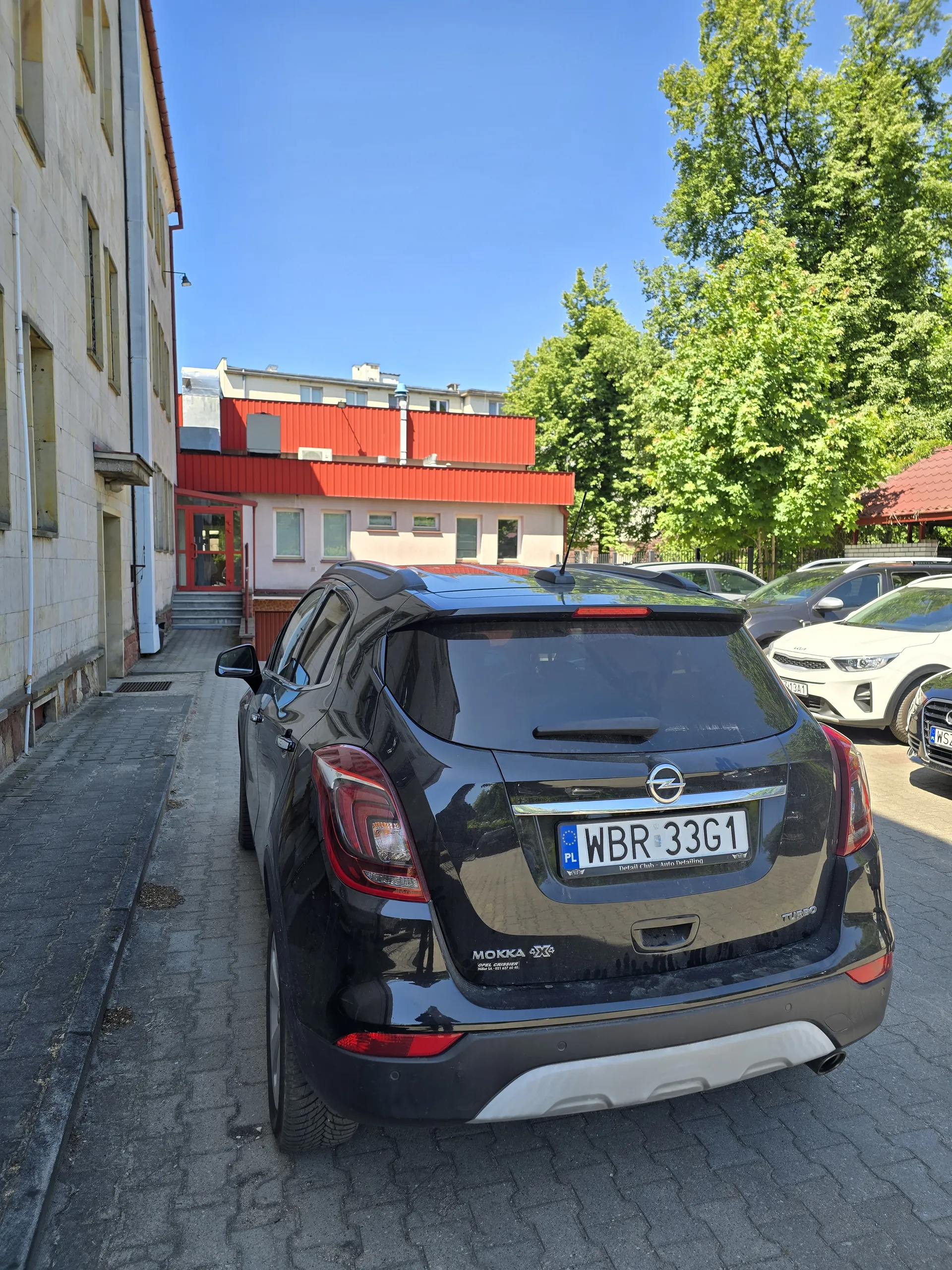 Czarny samochód marki Dacia z tablicą rejestracyjną WBR 33G1 zaparkowany na brukowanej powierzchni. W tle inne pojazdy, budynki z czerwonymi dachami, zielone drzewa i błękitne niebo bez chmur. Tablica rejestracyjna zawiera napis PL oraz numer WBR 33G1. Widoczne są również elementy architektoniczne budynków i brukowana droga.