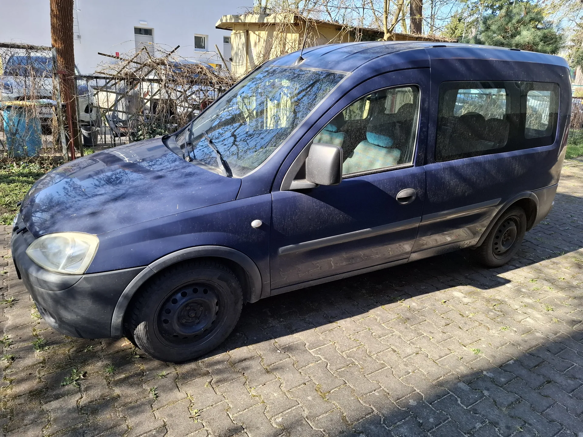 Ciemnoniebieski samochód dostawczy zaparkowany na brukowanej powierzchni. Pojazd ma czarne felgi, przyciemniane szyby i widoczne ślady brudu na karoserii. W tle budynek, suchy pnącza na konstrukcji, drzewa oraz niebieski pojemnik. Słońce rzuca cienie na bruk.