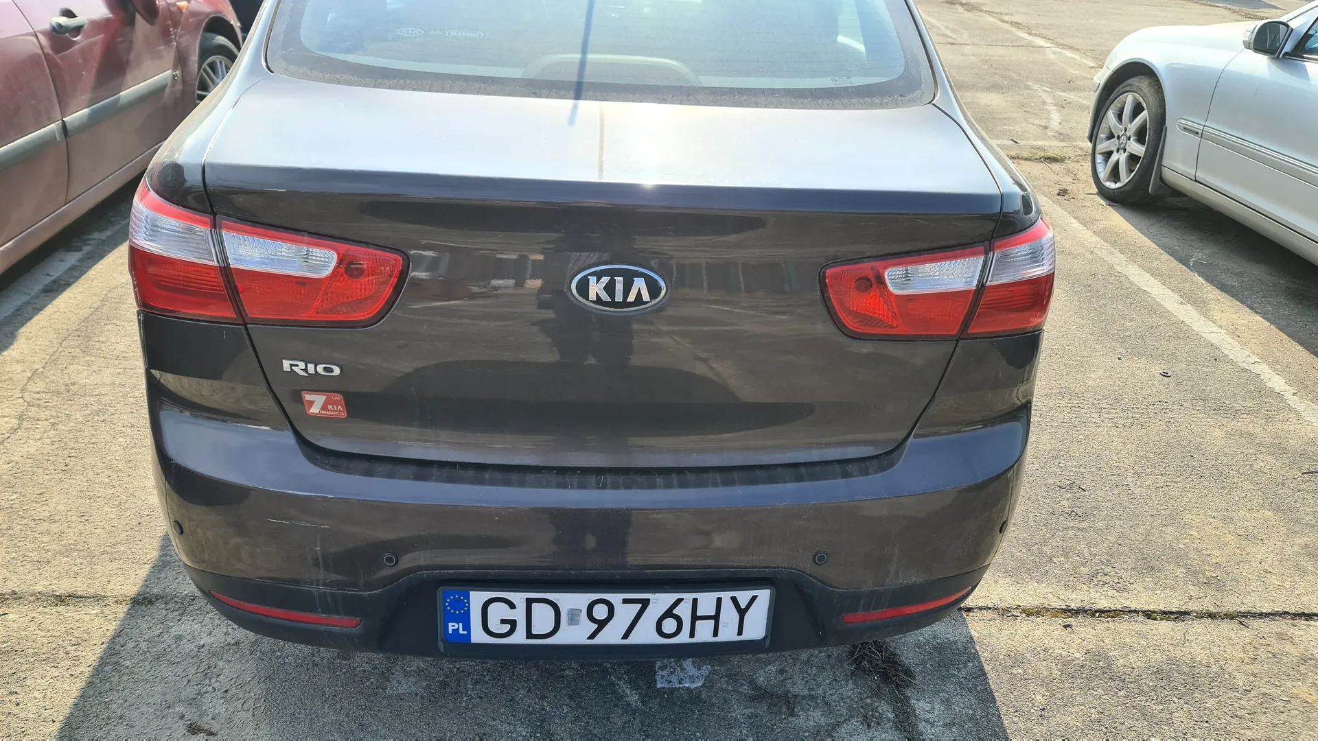 Tył ciemnoszarego samochodu marki KIA model Rio zaparkowanego na betonowym placu parkingowym. Na bagażniku widoczne logo KIA, napis "RIO" oraz czerwony naklejka z cyfrą "7". Tablica rejestracyjna z polskim kodem "PL" i numerem "GD 976HY". Po lewej stronie czerwony samochód, po prawej srebrny. Tło stanowi asfaltowa powierzchnia z białymi liniami parkingu.