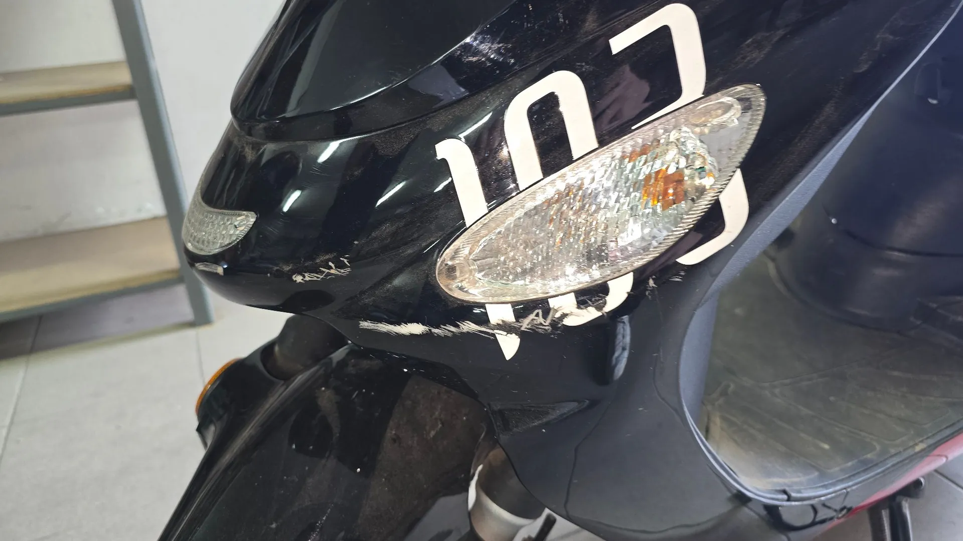 Zbliżenie na czarną, porysowaną obudowę skutera. Na powierzchni widoczny jest biały napis „103” oraz przezroczysty klosz kierunkowskas. Lakier w kilku miejscach jest przetarty i zarysowany.