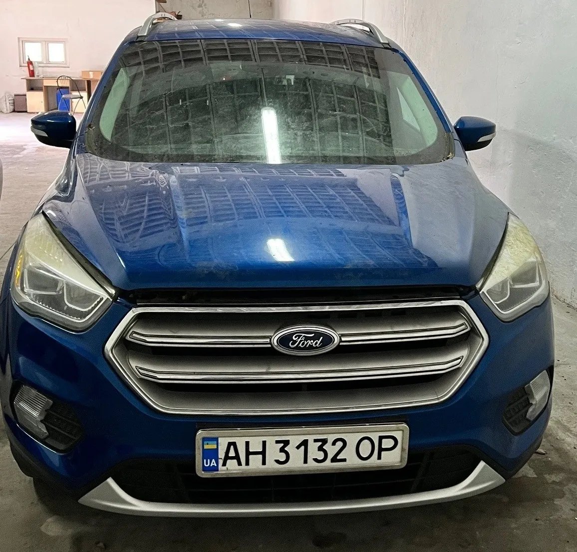 Niebieski samochód marki Ford z przodu, z widocznym logo na atrapie chłodnicy. Tablica rejestracyjna z napisem UA AH 3132 OP oraz flagą Ukrainy po lewej stronie. Samochód znajduje się w pomieszczeniu przypominającym garaż lub warsztat z betonowymi ścianami, oknami i meblami w tle. Odbicia na przedniej szybie i masku wskazują na oświetlenie z góry.