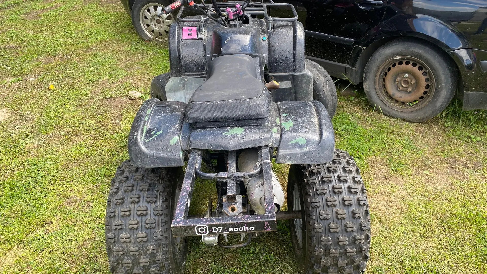 Widok z tyłu na czarny quad zaparkowany na trawie obok czarnego samochodu. Quad ma szerokie, terenowe opony z wyraźnym bieżnikiem, czarne błotniki z widocznymi zielonymi odpryskami farby oraz czarne siedzenie. Na tylnej ramie quada znajduje się naklejka z napisem 'D7_socha' oraz ikoną Instagrama.