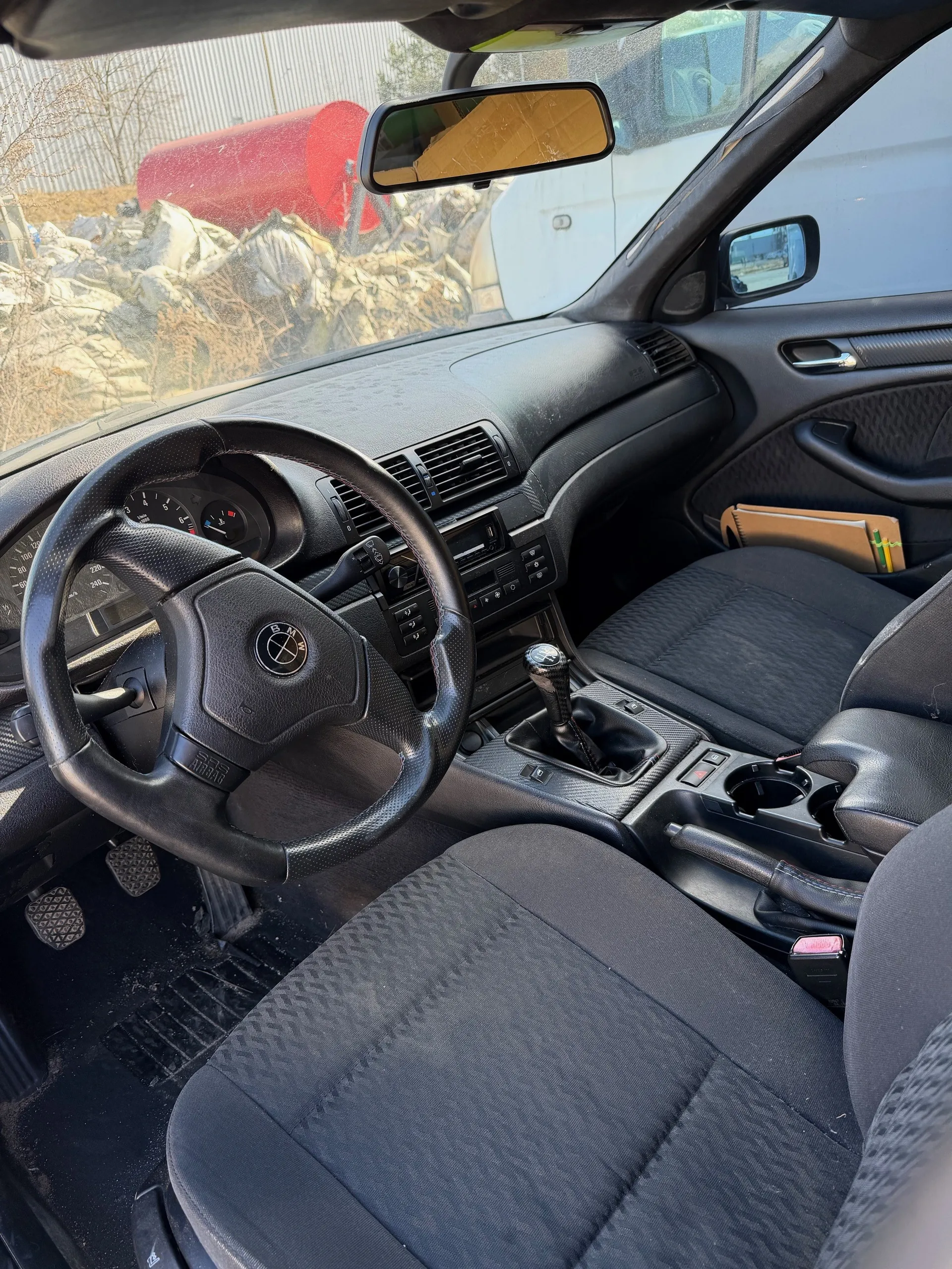 Wnętrze samochodu marki BMW z czarną kierownicą z logo, czarną tapicerką siedzeń, deską rozdzielczą z licznikami, dźwignią zmiany biegów i lusterkiem wstecznym. Przez przednią szybę widoczny czerwony cylinder i sterta materiałów na zewnątrz.