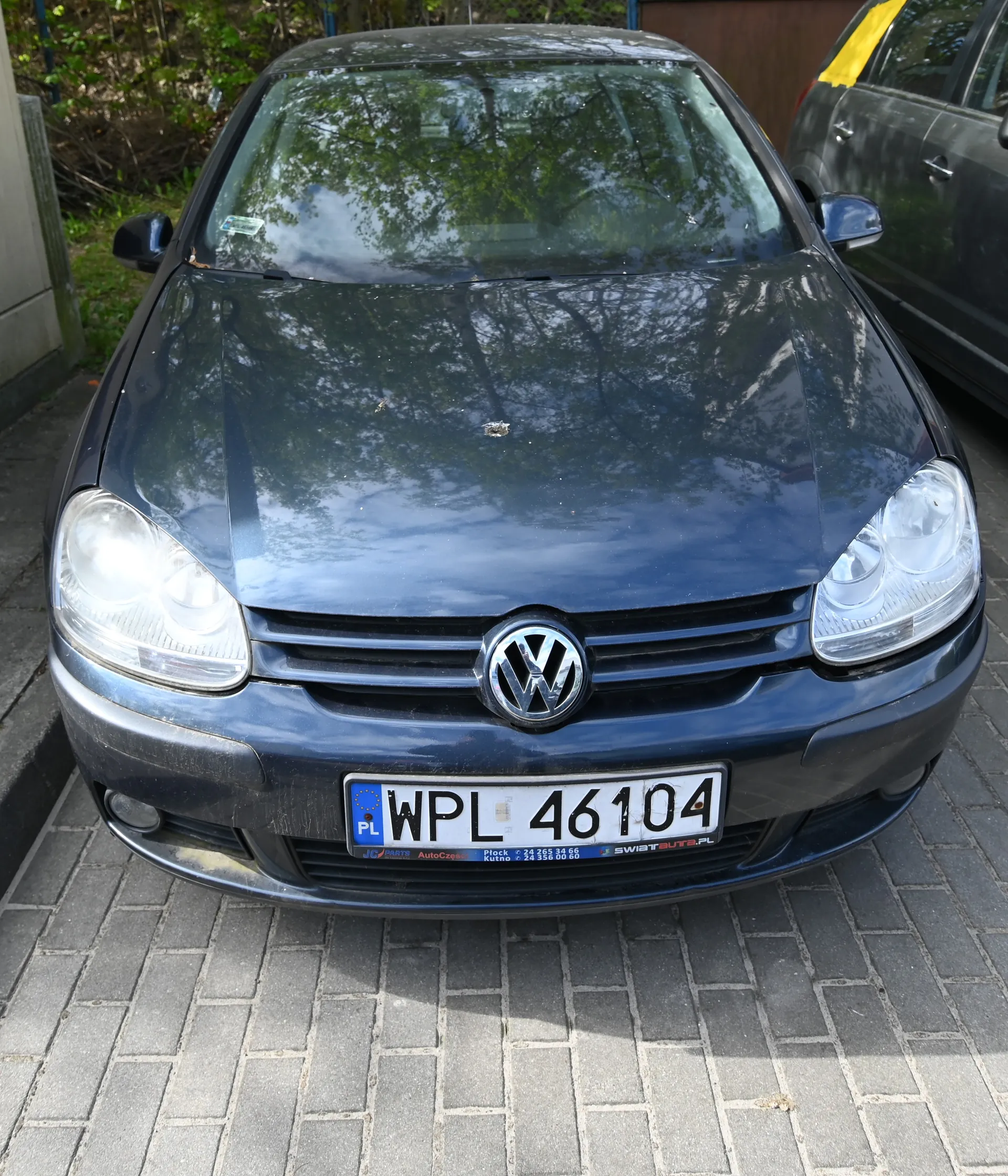 Ciemnoniebieski samochód marki Volkswagen z przodu, z tablicą rejestracyjną WPL 46104 (PL po lewej stronie). Samochód zaparkowany na brukowanej powierzchni, w tle częściowo widoczny inny pojazd z żółtą taśmą na dachu oraz zieleń i budynki. Logo VW na środku atrapy chłodnicy, reflektory włączone.