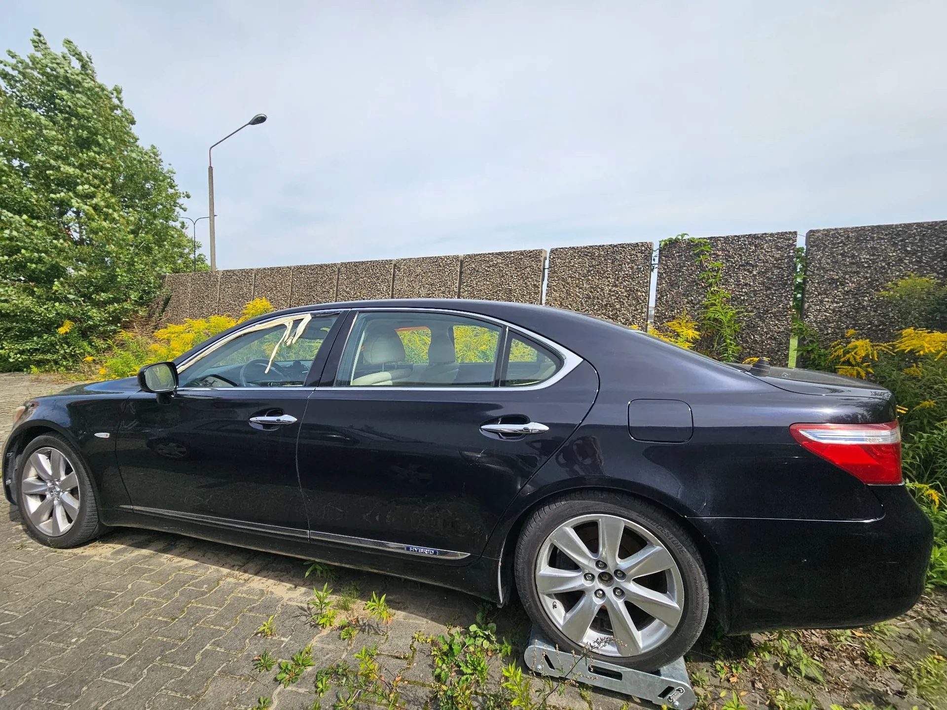 Czarny samochód osobowy typu sedan zaparkowany bokiem na kostce brukowej. Pojazd posiada srebrne felgi aluminiowe i napis 'HYBRID' na dolnej części drzwi. W tle widoczny jest wysoki mur oporowy z gabionów wypełnionych kamieniami, zielone krzewy, żółte kwiaty oraz latarnia uliczna pod jasnym, zachmurzonym niebem.