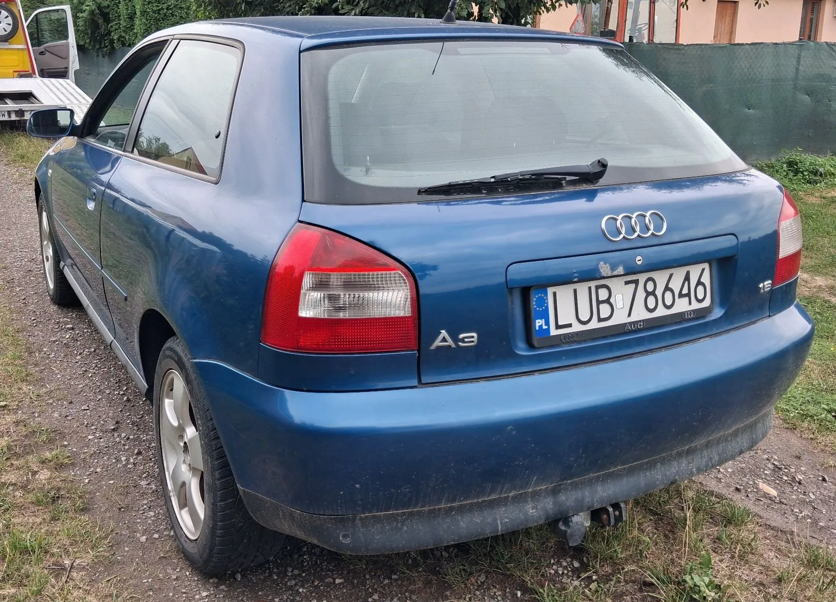 Niebieski samochód marki Audi A3 zaparkowany na żwirowej drodze. Widok z tyłu i z boku. Samochód posiada polską tablicę rejestracyjną o numerze LUB 78646. Na klapie bagażnika widoczne jest logo Audi oraz oznaczenie modelu A3.