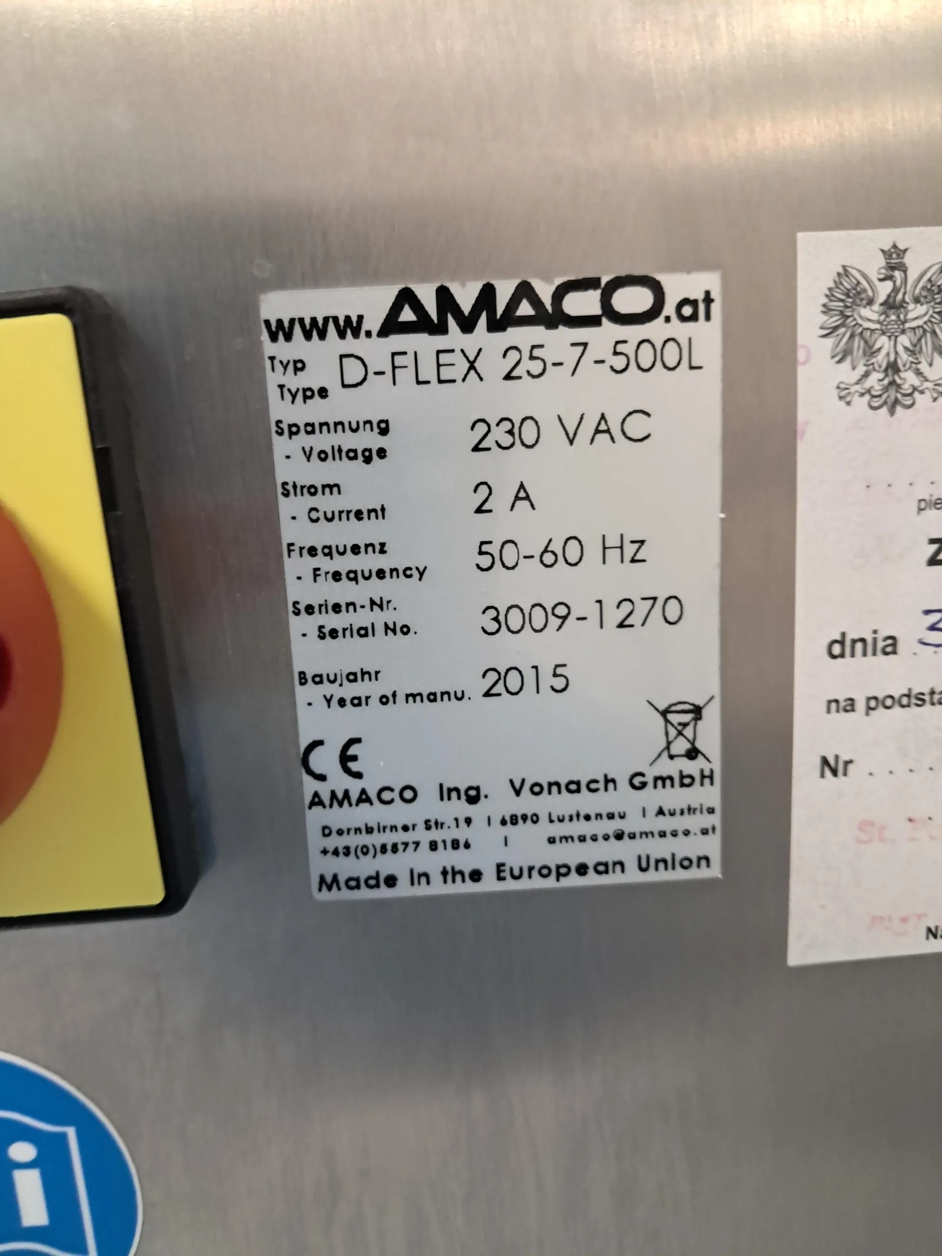 Srebrna tabliczka znamionowa przymocowana do metalowej powierzchni. Zawiera następujący tekst: 'www.AMACO.at', 'Typ D-FLEX 25-7-500L', 'Spannung - Voltage 230 VAC', 'Strom - Current 2 A', 'Frequenz - Frequency 50-60 Hz', 'Serien-Nr. - Serial No. 3009-1270', 'Baujahr - Year of manu. 2015'. Poniżej znajduje się znak CE oraz dane producenta: 'AMACO Ing. Vonach GmbH, Dornbirner Str.19 | 6890 Lustenau | Austria, +43(0)5577 8186 | amaco@amaco.at' oraz napis 'Made in the European Union'. Obok tabliczki widoczny jest fragment białej kartki z godłem Polski.