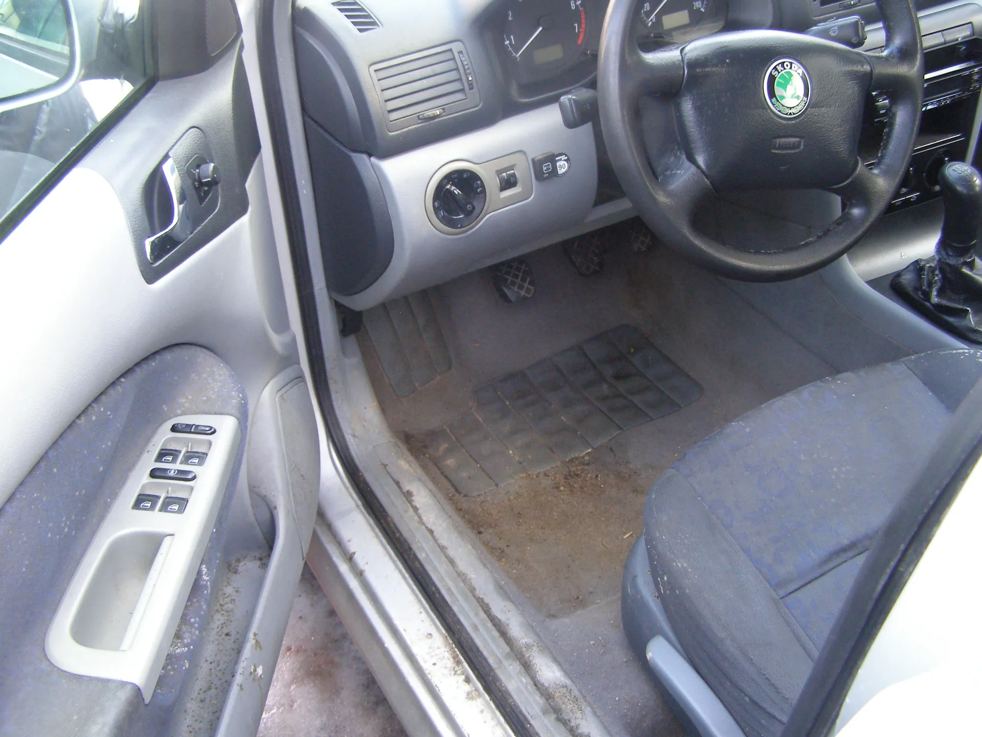 Wnętrze samochodu z otwartą drzwiami kierowcy. Widoczna kierownica z logo Škoda, deska rozdzielcza z licznikami, gałka zmiany biegów, siedzenie kierowcy oraz panel drzwiowy z przyciskami sterowania szybami. Podłoga i próg drzwi pokryte są brudem, a dywaniki mają siatkowy wzór. Wnętrze posiada szare i czarne elementy wykończeniowe.