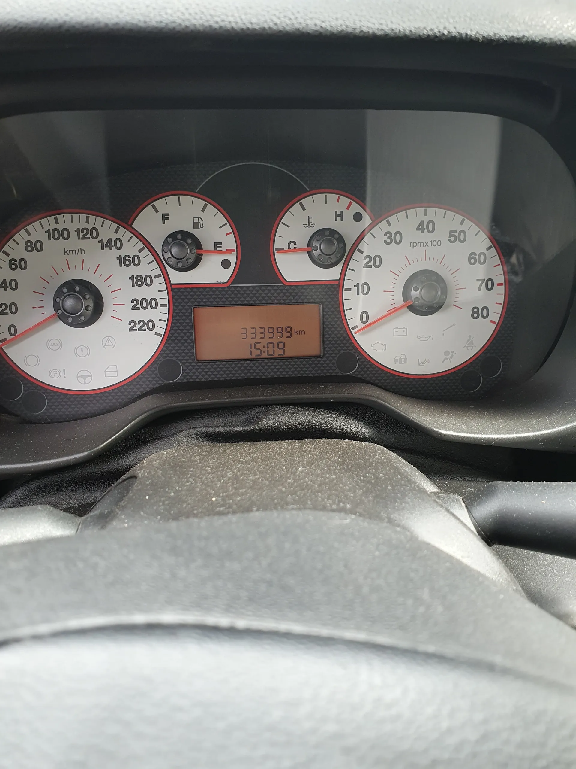 Tablica przyrządów samochodowych z licznikiem prędkości (0-220 km/h), tachometrem (0-80 x100 obr./min), wskaźnikiem temperatury chłodniczego (H/C), wskaźnikiem paliwa (F/E) oraz wyświetlaczem cyfrowym pokazującym przebieg 333999 km i godzinę 15:09. Widoczne są również różne lampki ostrzegawcze.