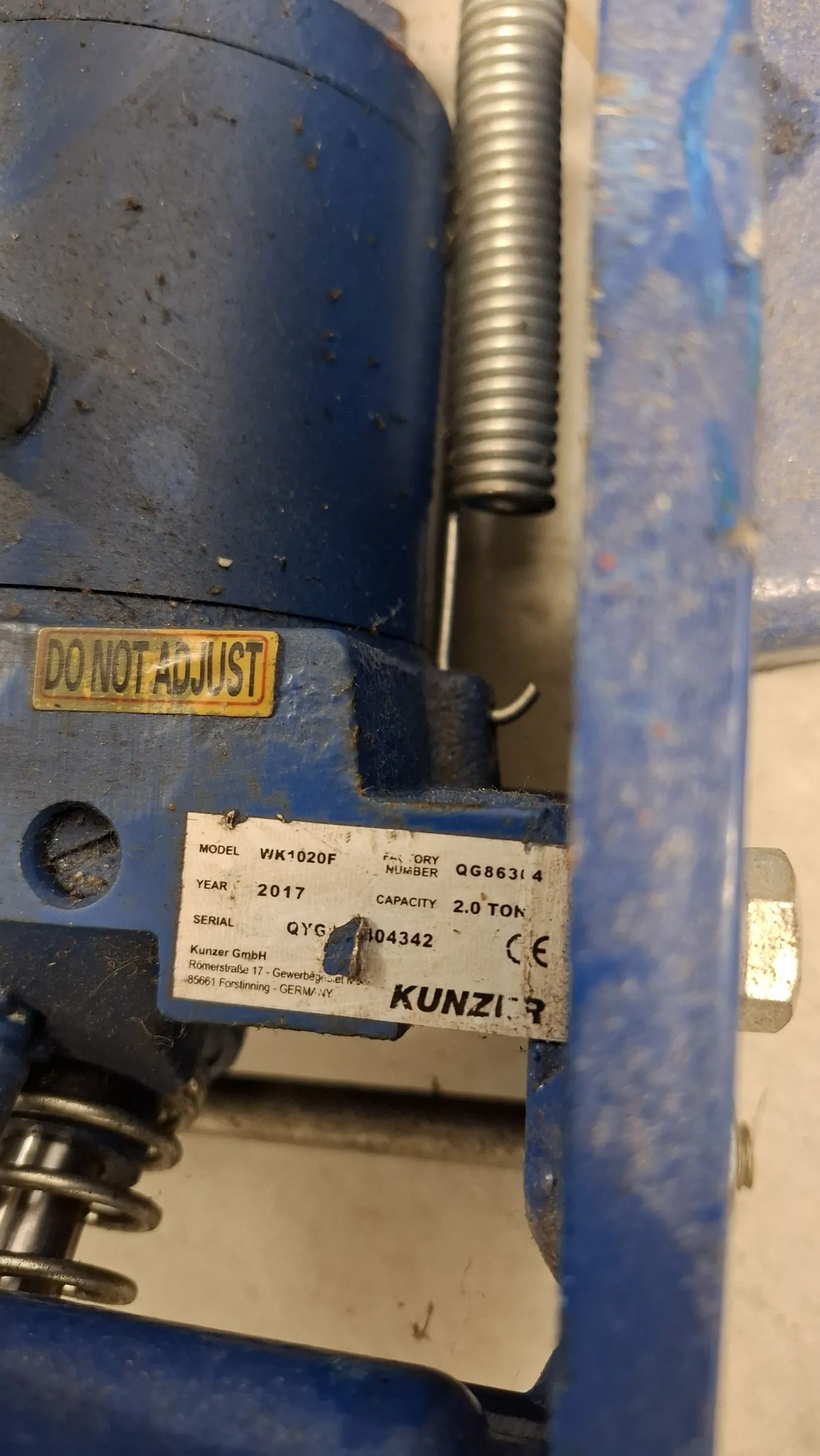 Zbliżenie na niebieską, metalową maszynę przemysłową. Na obudowie znajduje się żółta naklejka z napisem „DO NOT ADJUST” oraz srebrna tabliczka znamionowa z tekstem: „MODEL WK1020F”, „FACTORY NUMBER QG86314”, „YEAR 2017”, „CAPACITY 2.0 TON”, „SERIAL QYG 104342”, logo CE oraz napis „Kunzer GmbH, Römerstraße 17 - Gewerbegebiet, 85061 Forstinning - GERMANY” i duży napis „KUNZIER”. Widoczna jest również metalowa sprężyna.