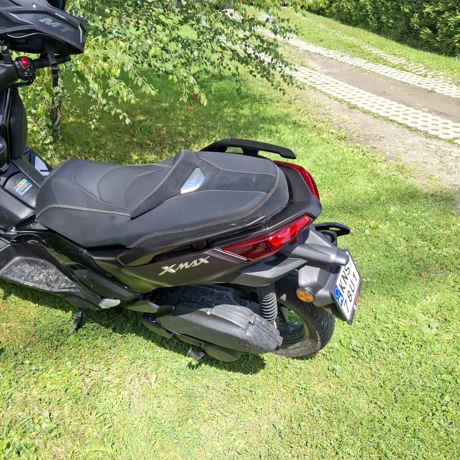 Czarny skuter marki Yamaha XMAX zaparkowany na trawie. Widoczny jest tył pojazdu z siedzeniem, tylną lampą, napisem 'XMAX' na boku oraz tablicą rejestracyjną o numerze 'S KNS 18U1'. W tle widać zielone drzewa i fragment betonowej drogi.