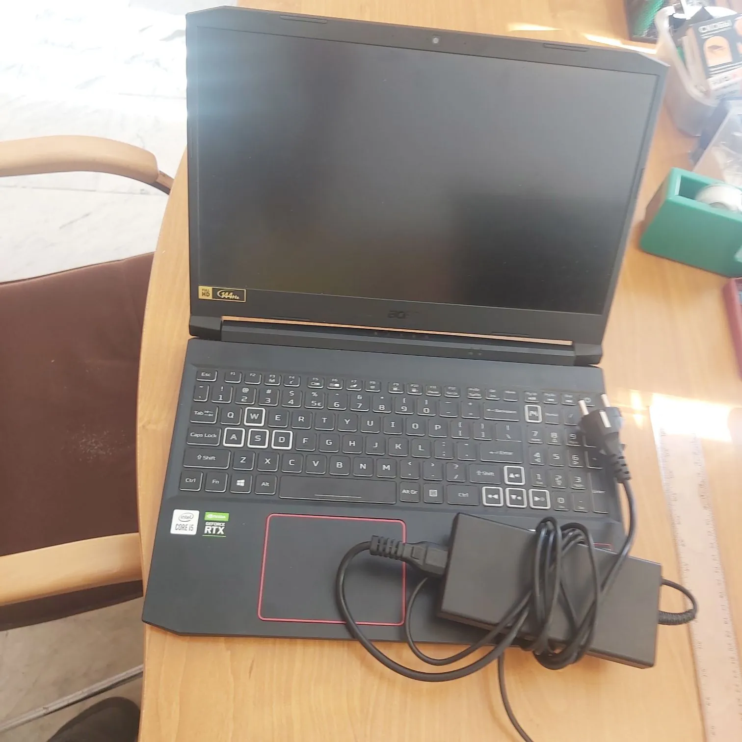Czarny laptop marki Acer z otwartym ekranem, leżący na drewnianym stole. Na obudowie widoczne są naklejki: złota naklejka 'Intel Core i7' oraz naklejka 'NVIDIA GeForce RTX'. Do laptopa podłączony jest zasilacz, który leży na klawiaturze i touchpadu.