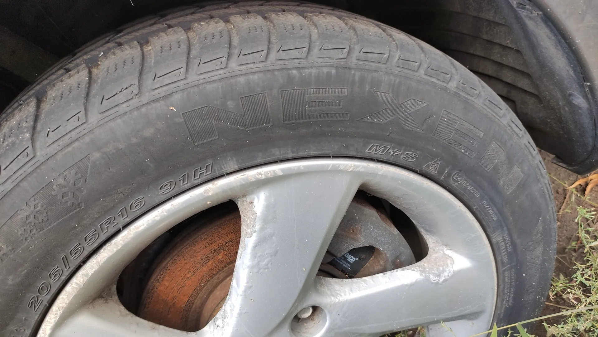 Zbliżenie na oponę samochodową marki Nexen. Na boku opony widoczny jest napis 'NEXEN', oznaczenie 'M+S' oraz rozmiar '205/55R16 91H'. Opona jest zamontowana na srebrnej, pięcioramiennej aluminiowej feldze, która wykazuje oznaki zużycia i zarysowań. Wewnątrz felgi widoczna jest zardzewiała tarcza hamulcowa.