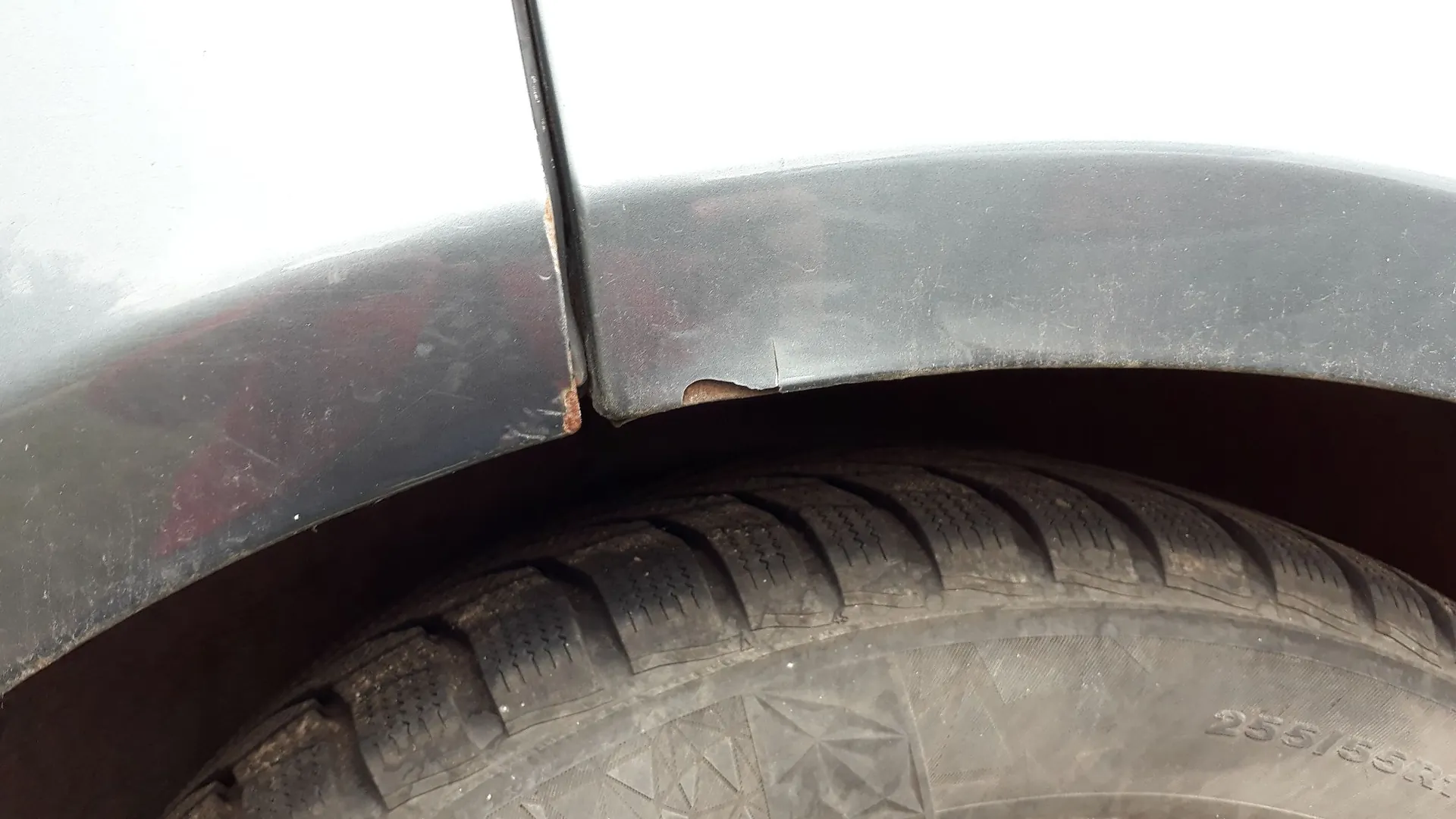 Zbliżenie na nadkole srebrnego samochodu. Widoczne są uszkodzenia lakieru, wgniecenie oraz ogniska korozji na krawędzi błotnika nad oponą. Na oponie widoczny jest fragment oznaczenia rozmiaru: 255/55R