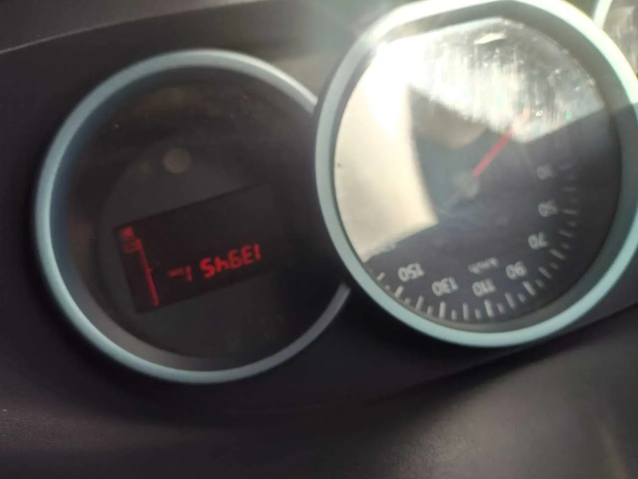 Zbliżenie na wskaźnik prędkości i licznik przebiegu w samochodzie. Wskaźnik prędkości z zakresem od 0 do 150 km/h, igła wskazująca blisko 0. Poniżej wyświetlacz cyfrowy z czerwonym tekstem '13945 km'.